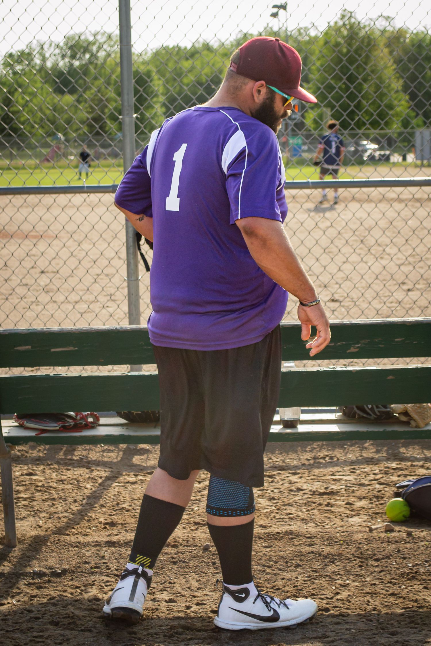 Male softball player wearing purple number 1 jersey and Nike shoes standing in the dugout area.