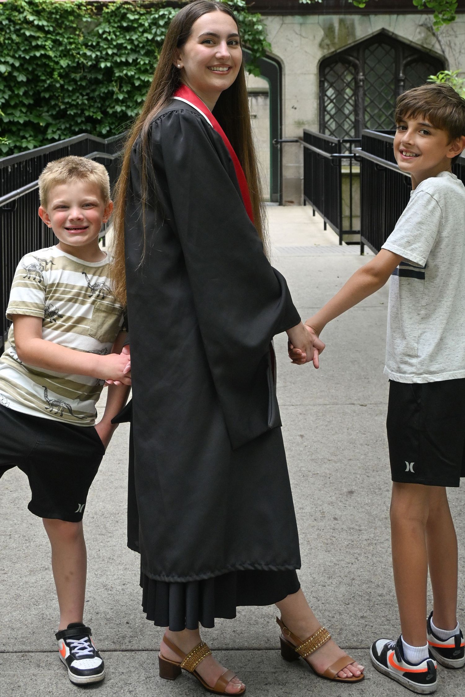 A smiling graduate in black cap and gown holds hands with two young boys outside a ivy-covered building.