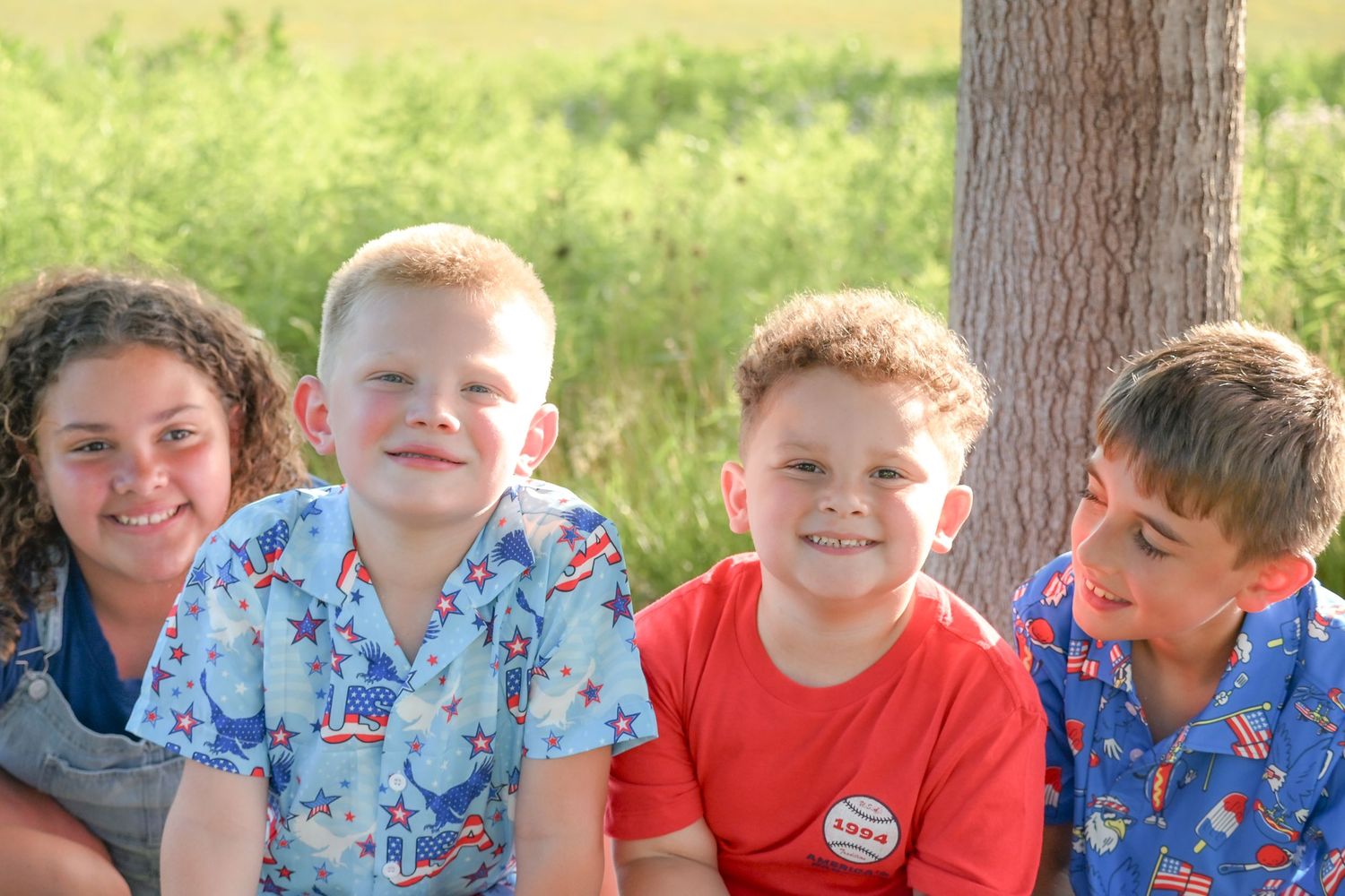 Four smiling children in patriotic outfits sitting together outdoors near a tree on a sunny day.