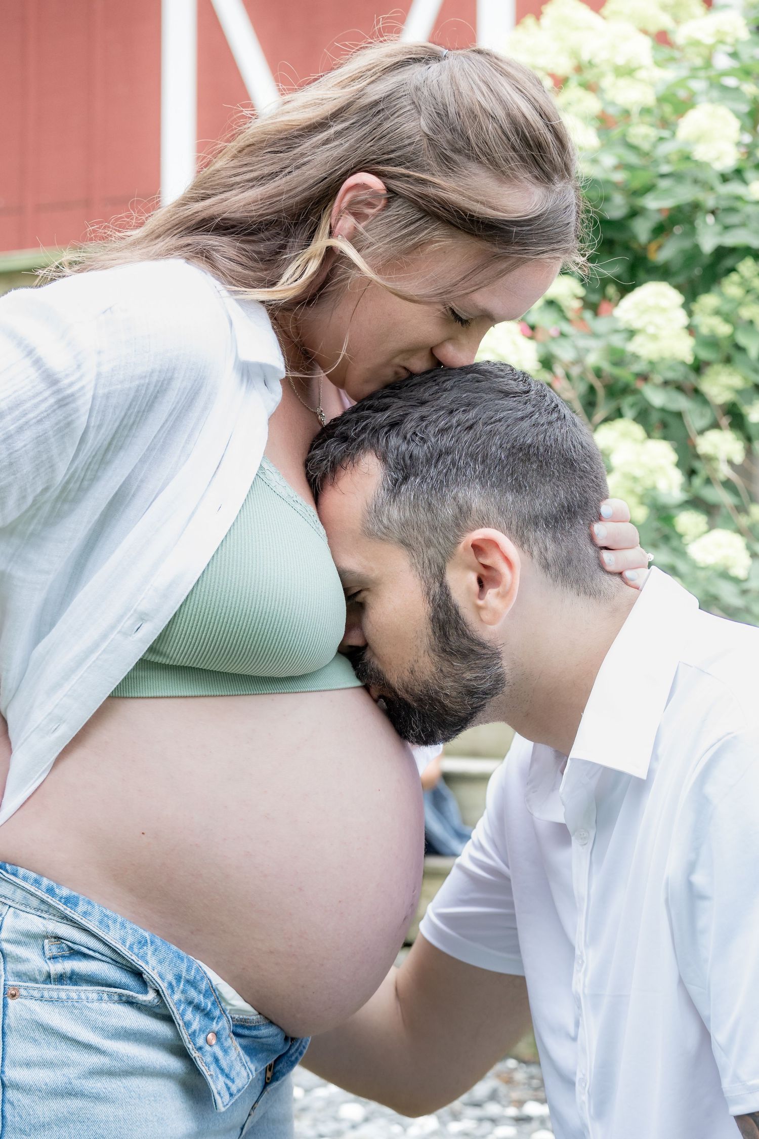 Expectant couple sharing tender moment as man kisses pregnant woman's baby bump in garden setting.