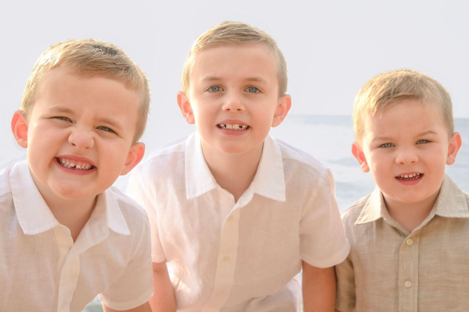 Three smiling boys in white shirts pose together on a bright beach for a cheerful family portrait photo.