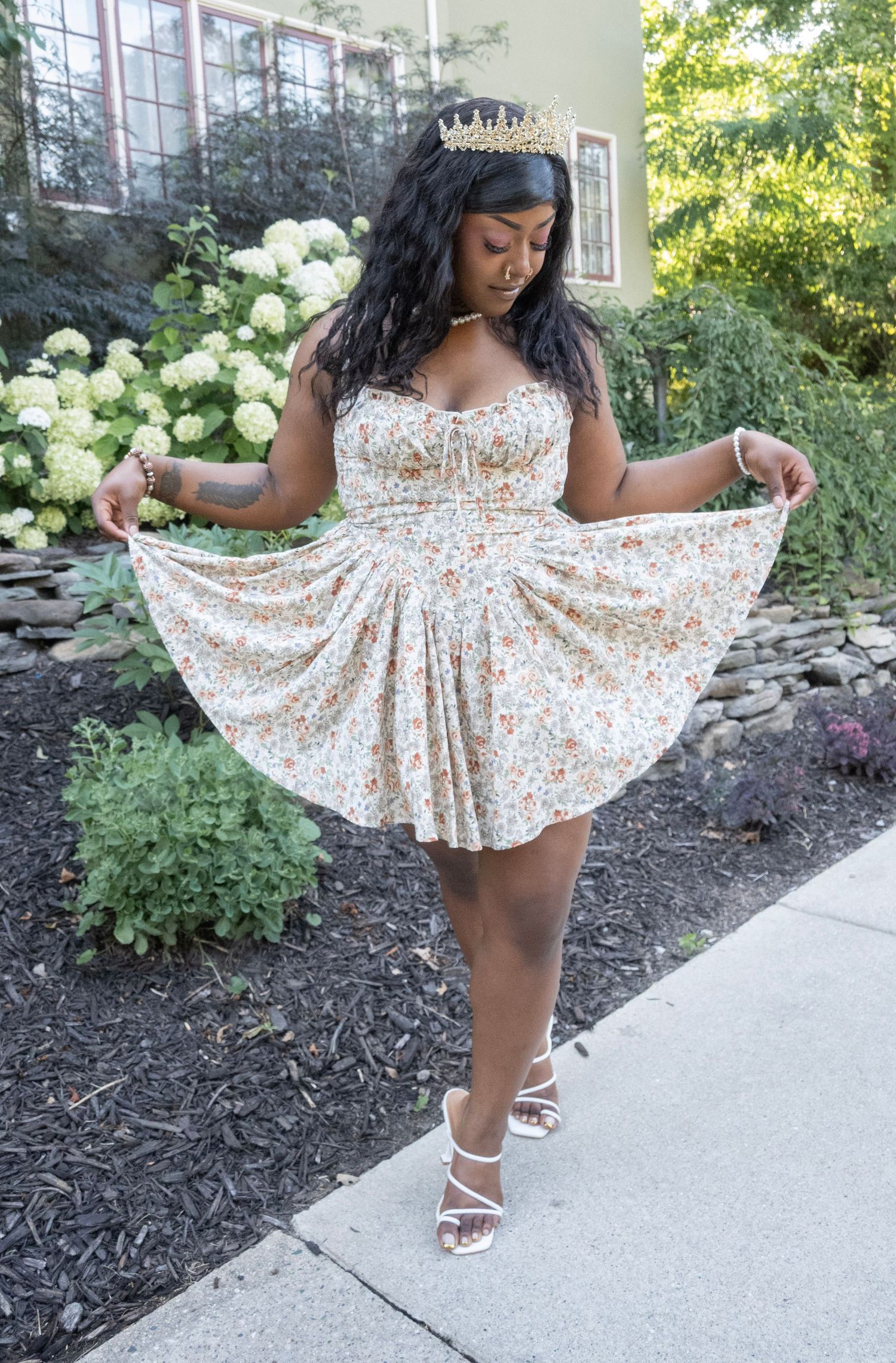 Woman in floral mini dress and gold crown poses outdoors near white hydrangeas, holding skirt wide.