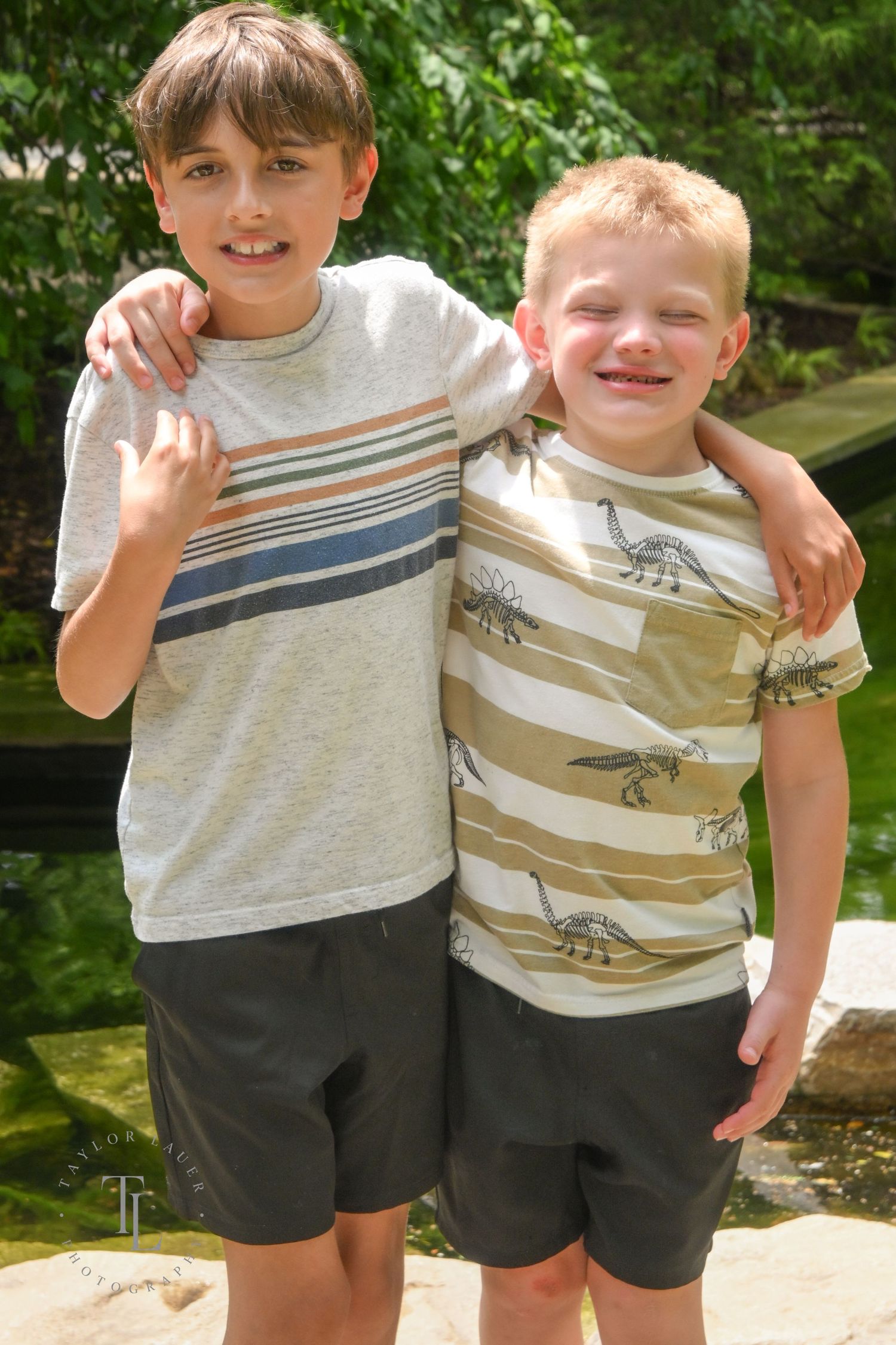 Two smiling boys in matching striped shirts and dark shorts pose with arms around each other outdoors.