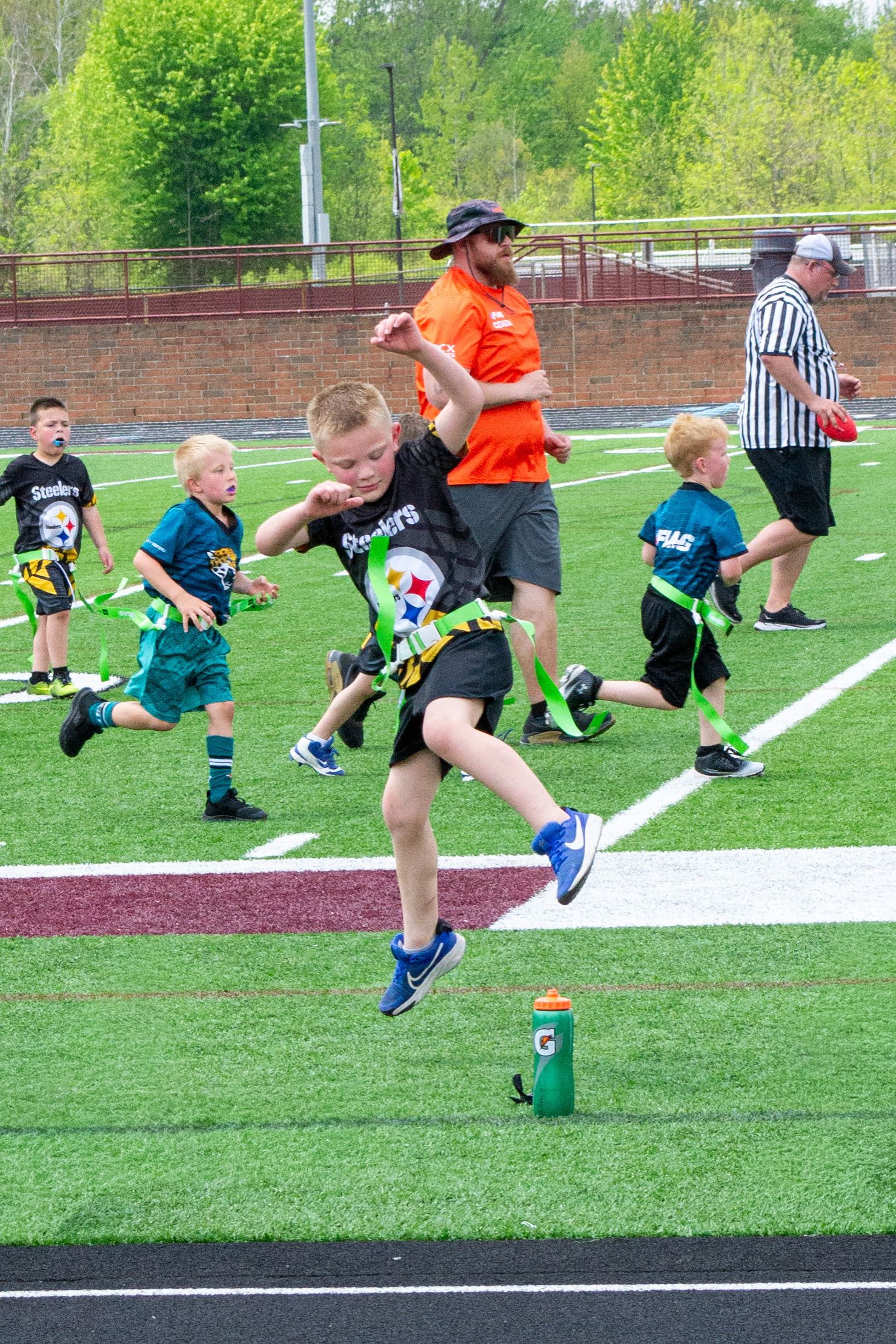 Kids playing youth flag football on turf field with coaches and referee supervising.