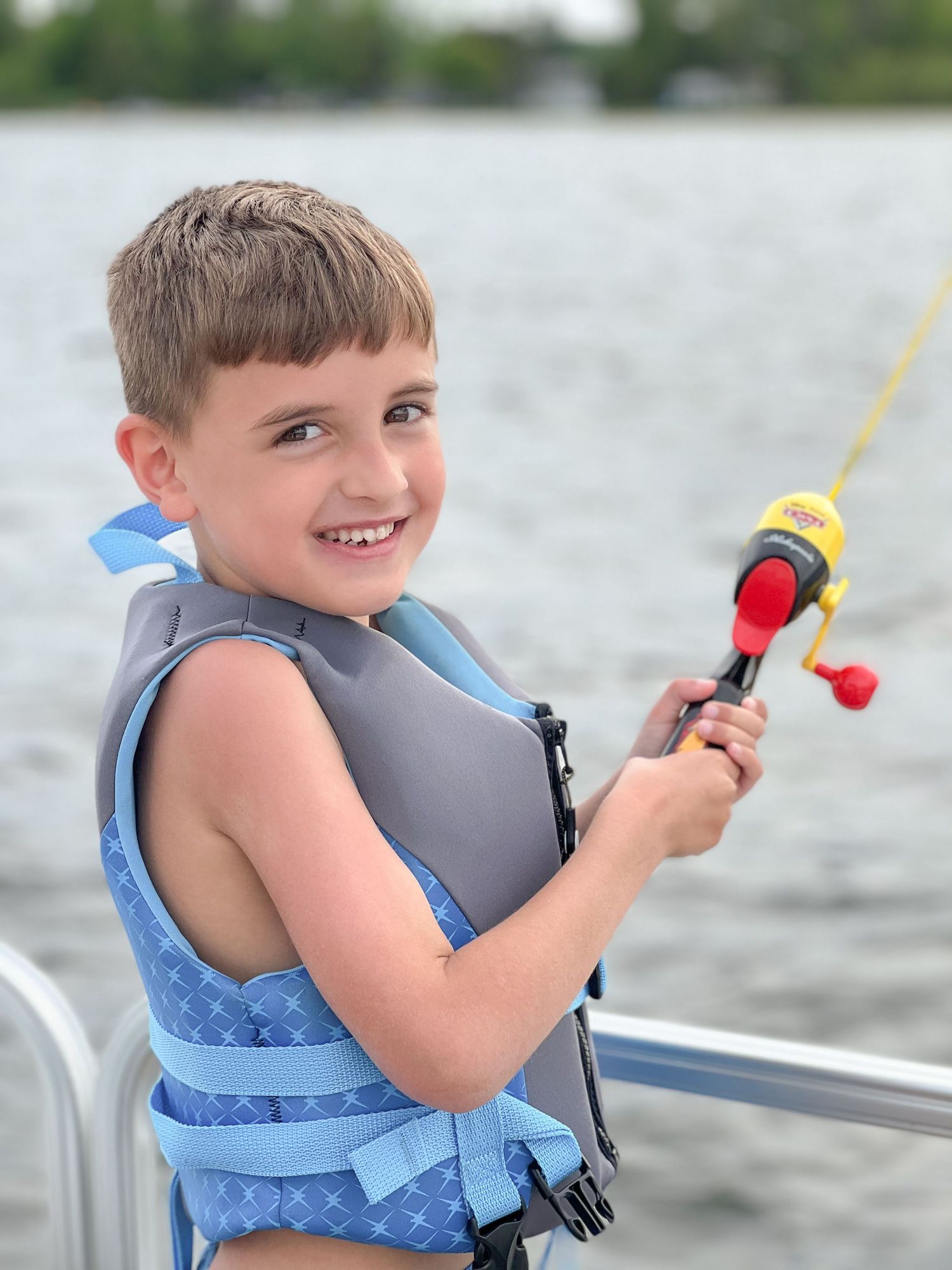 Smiling boy in life jacket holding a fishing rod on a boat on the lake.