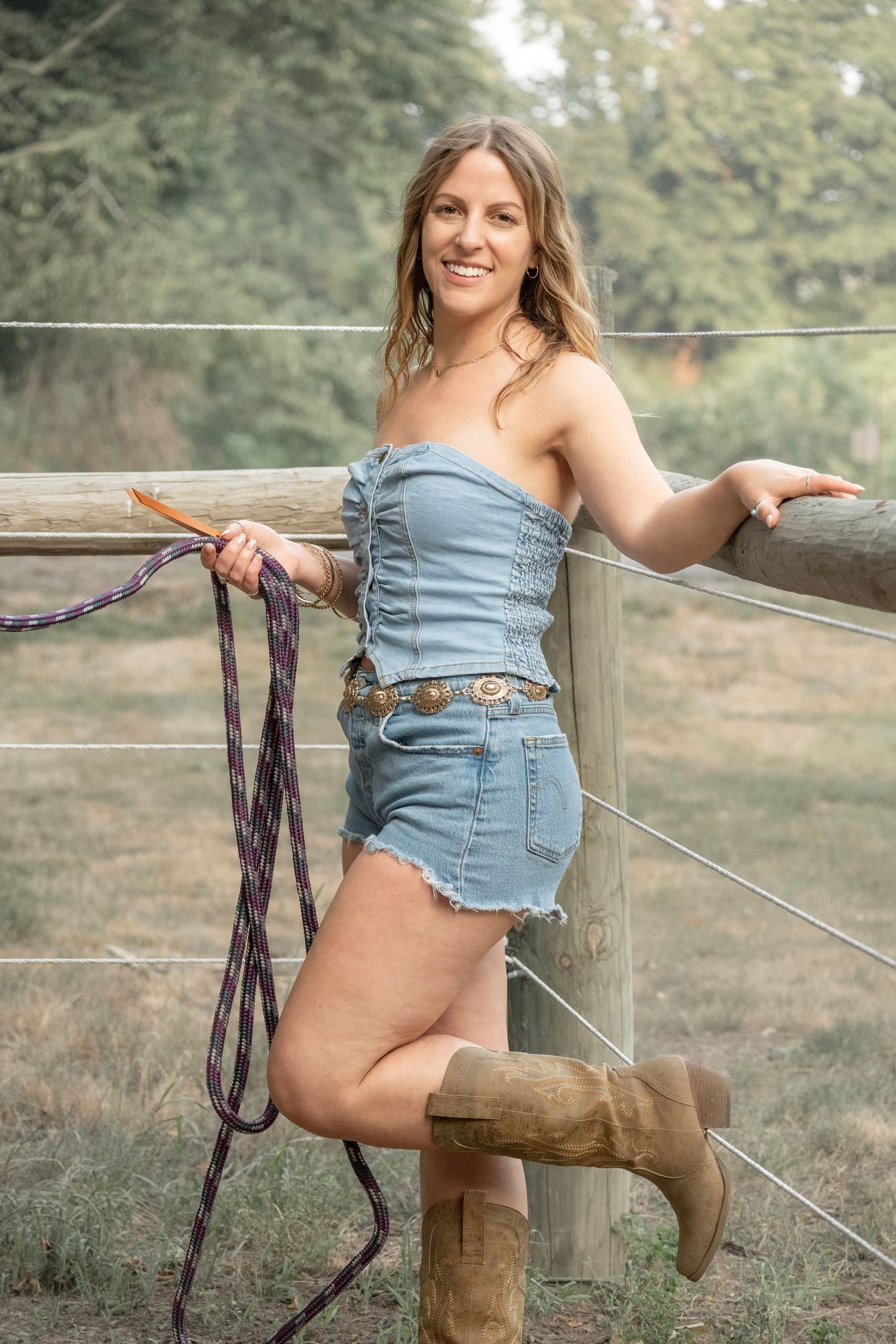 Woman in denim romper and cowboy boots leaning on wooden fence post holding horse lead rope on a farm.
