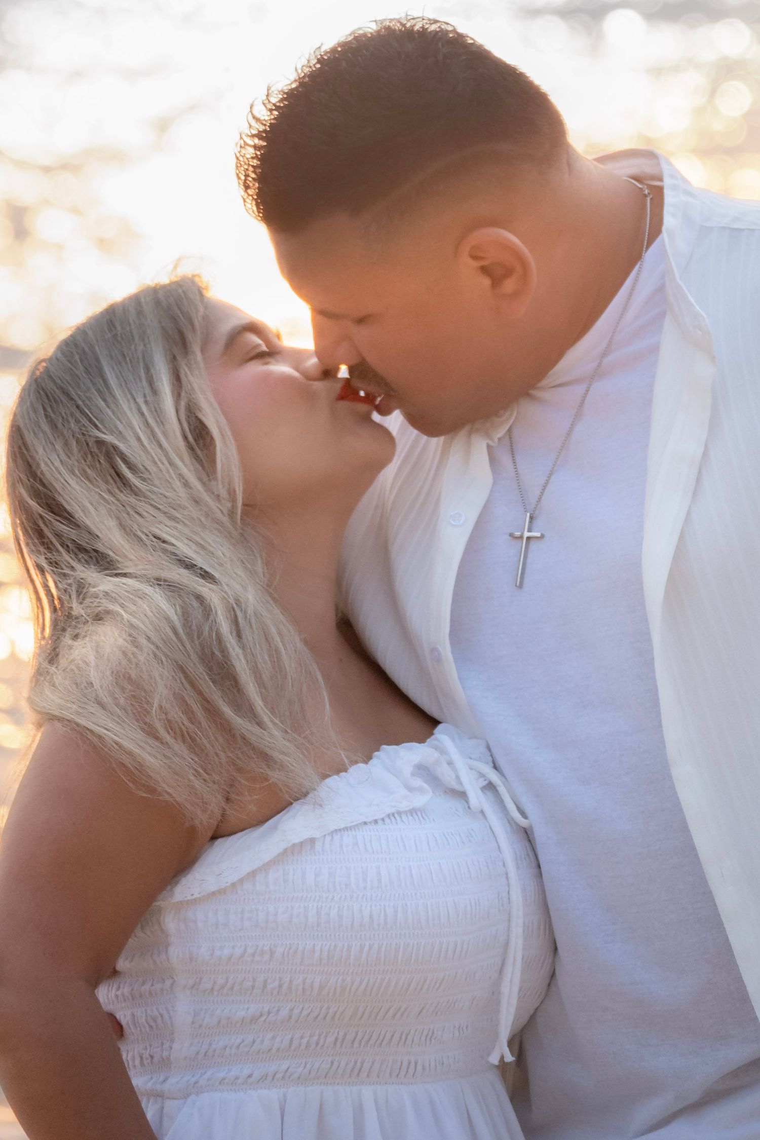 Couple sharing a romantic kiss at sunset on the beach, both dressed in white, man wearing a cross necklace.