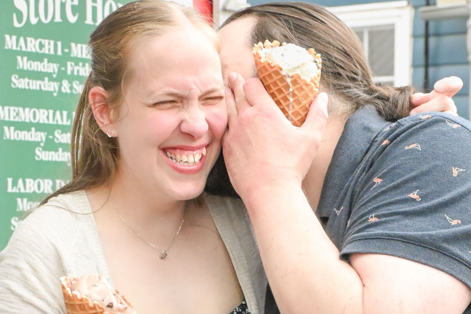 A laughing couple playfully pressing ice cream cones together outside an ice cream store.