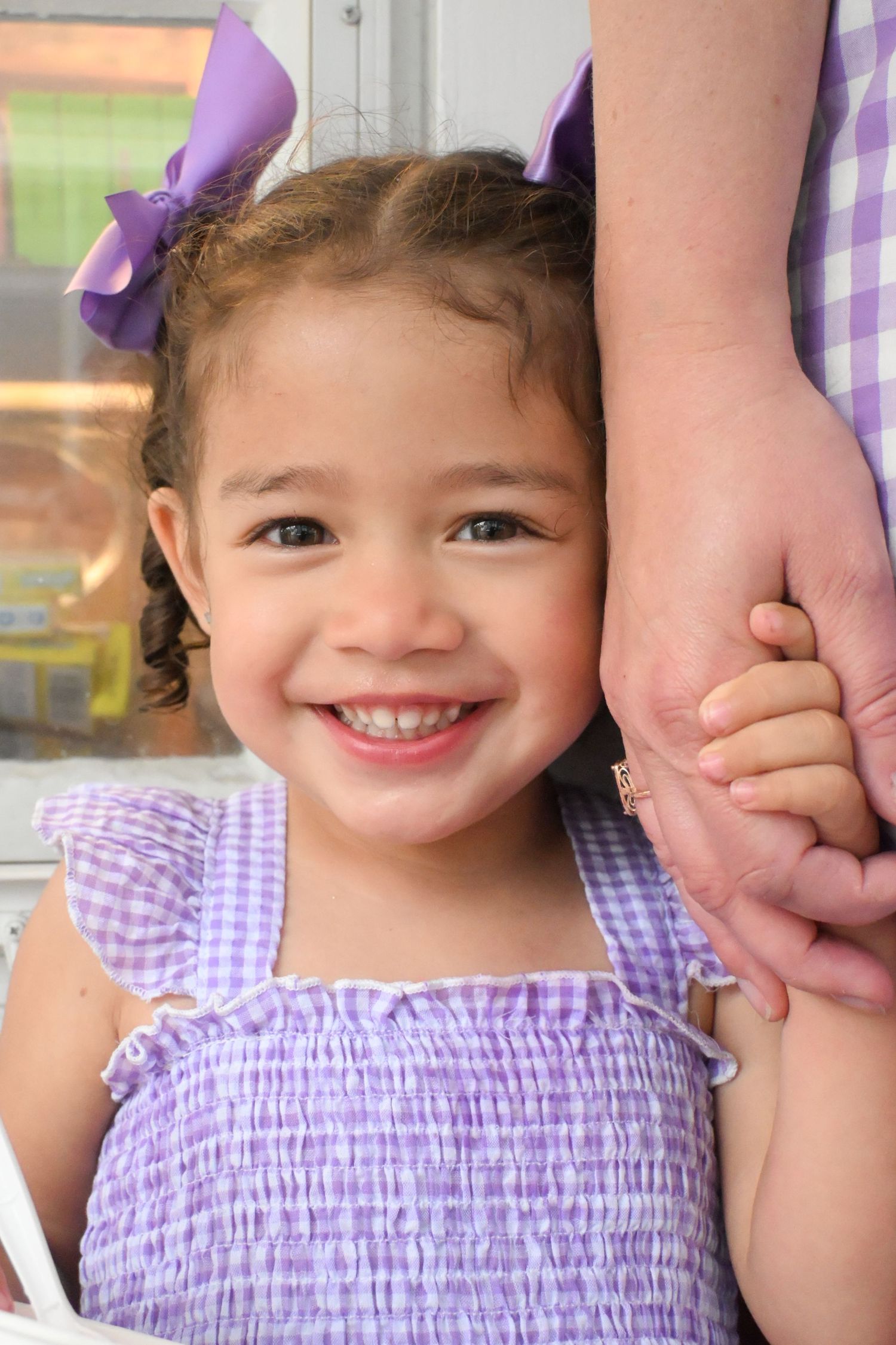 A cheerful toddler girl in a purple gingham dress with purple bows smiles while holding an adults hand.