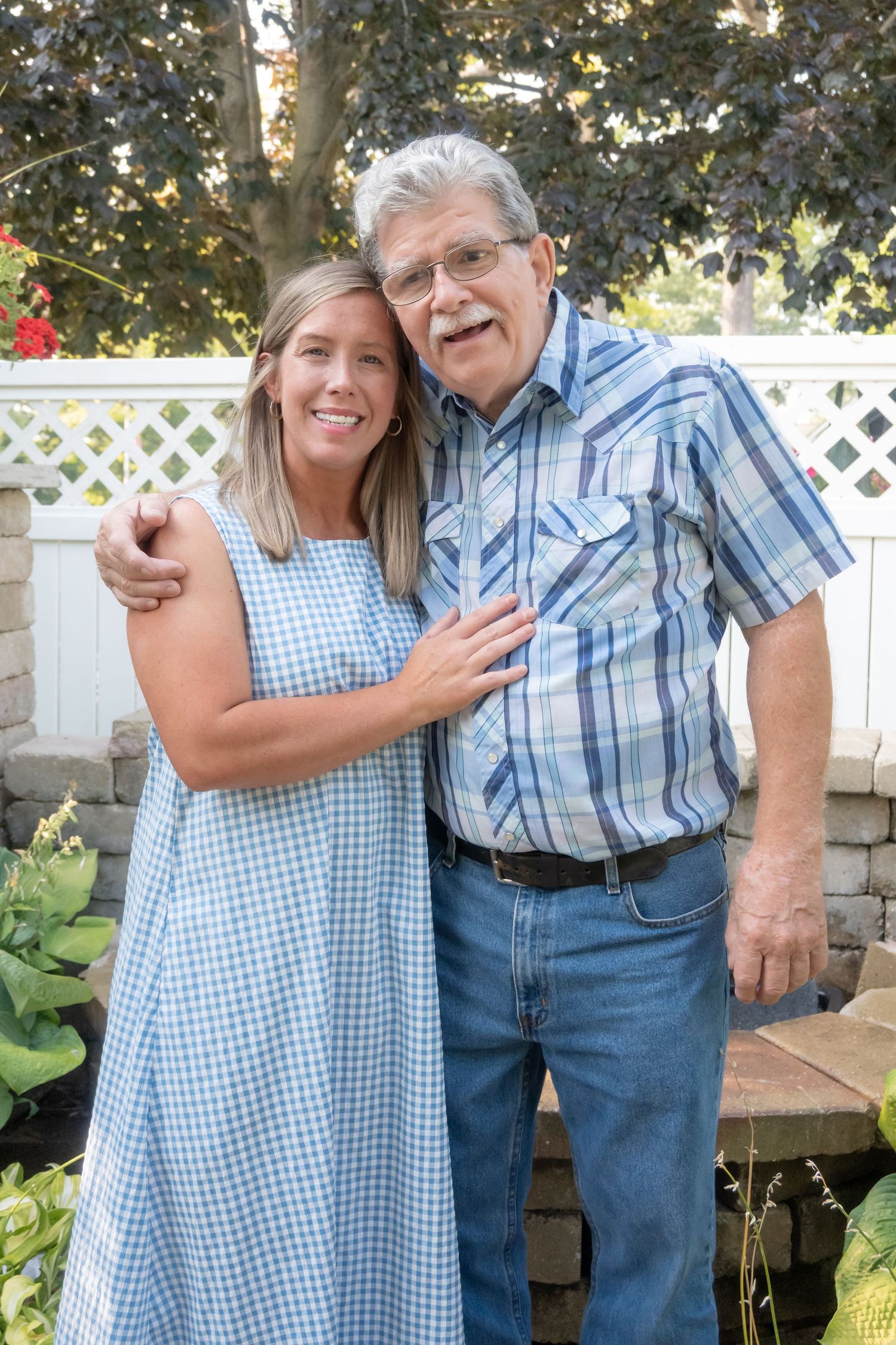 An older man and younger woman smile together in a garden, wearing blue outfits, posing in front of a white lattice fence.