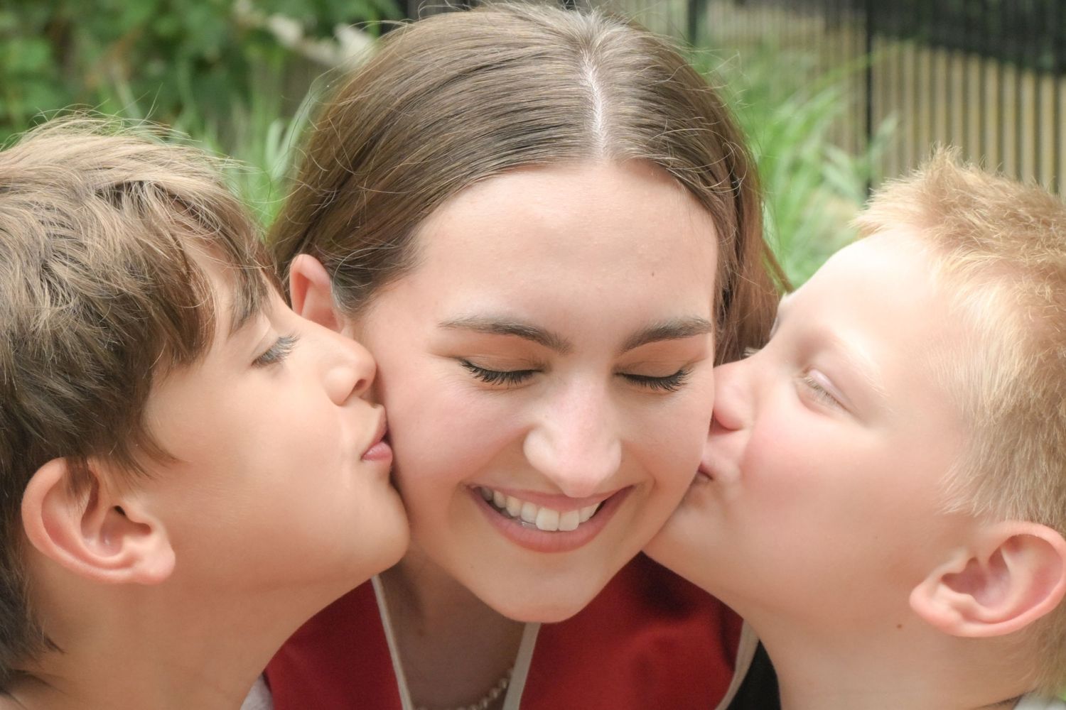 Two boys kissing a smiling young woman on each cheek outdoors.