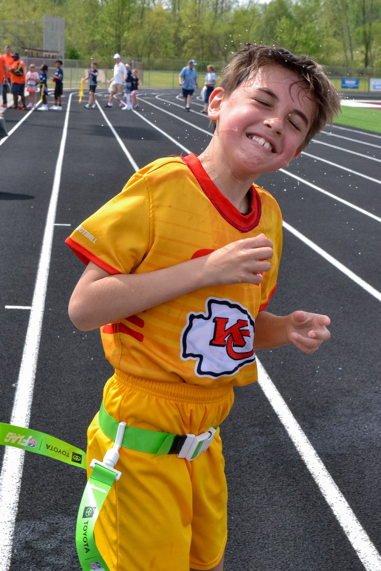 Smiling boy in Kansas City Chiefs yellow jersey running on a black track during a flag football game.