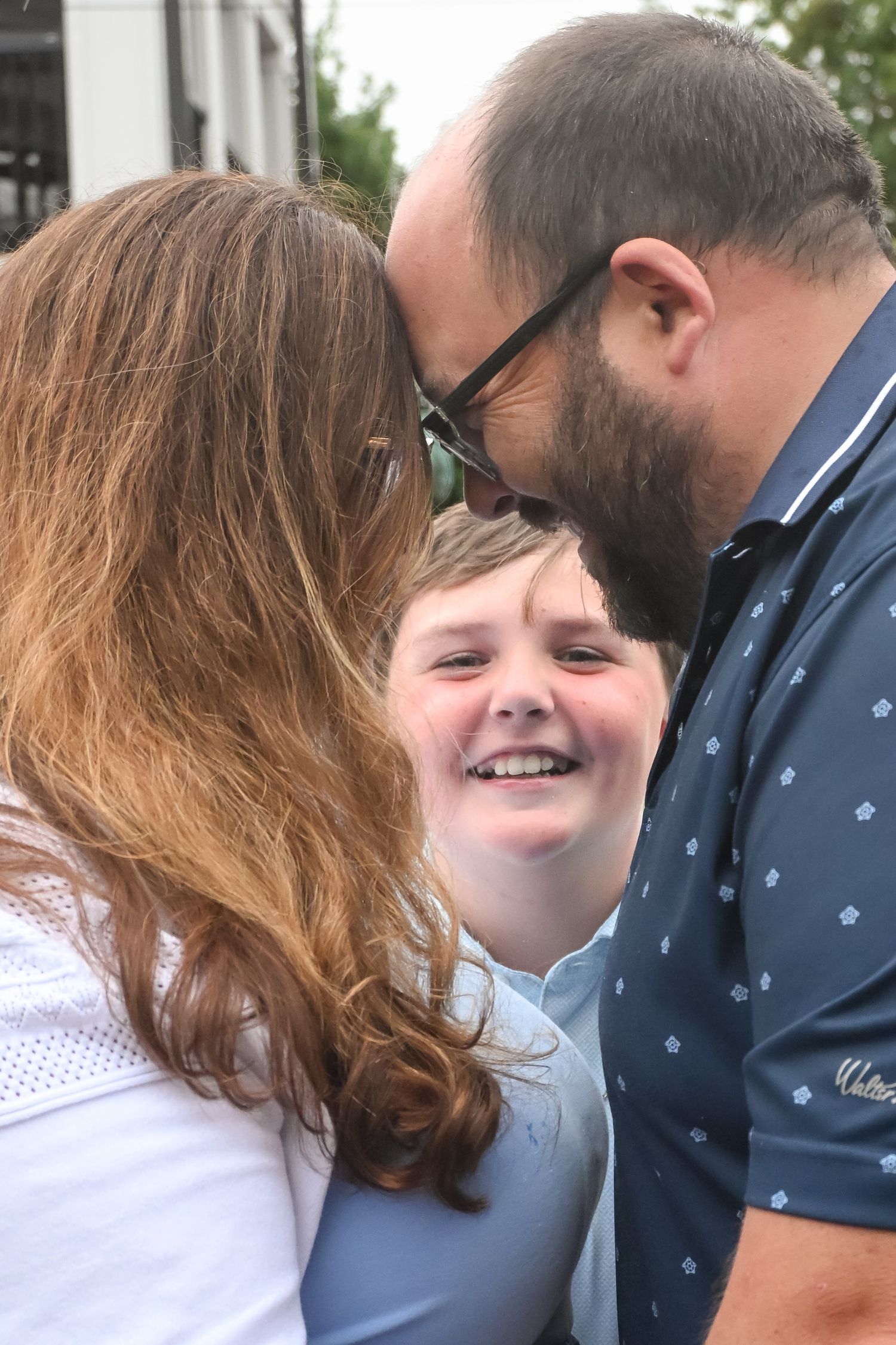 A joyful boy smiles as his parents lean in to kiss his cheeks outdoors.