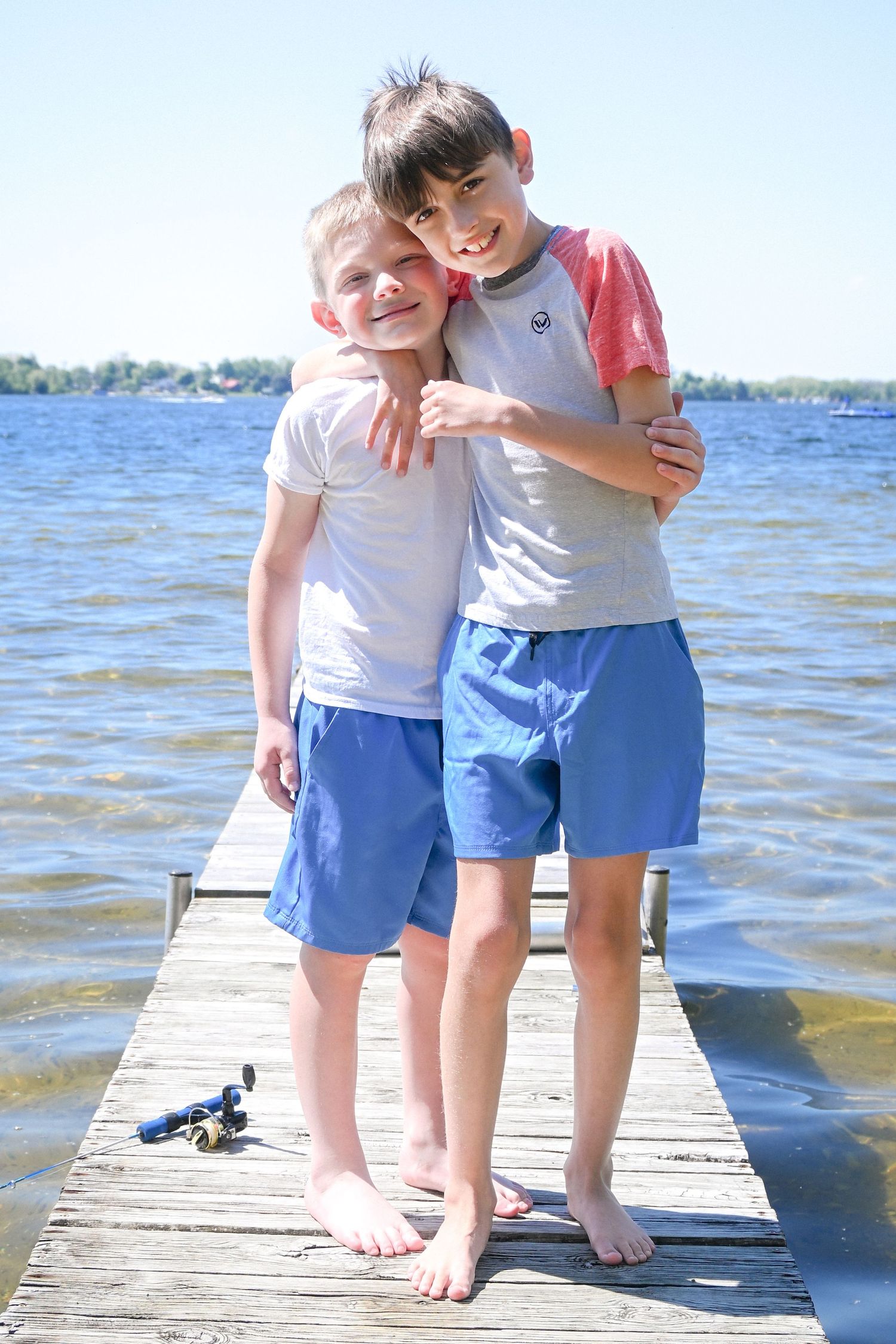 Two smiling boys hugging on a wooden dock by a lake on a sunny day, wearing blue shorts and t-shirts.