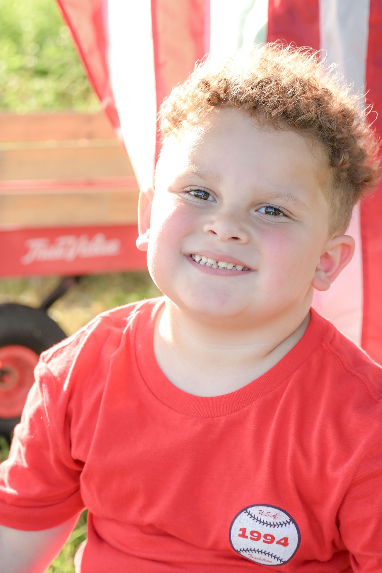 Smiling red-haired boy in a red 1994 baseball shirt sitting near a red wagon outdoors.