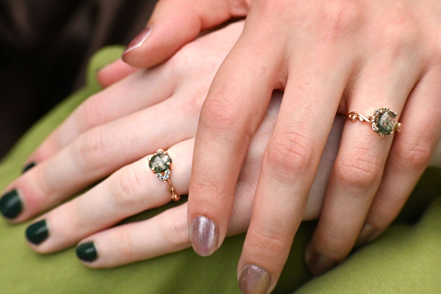 Two hands wearing matching gold rings with green gemstones, one hand with dark green nail polish.