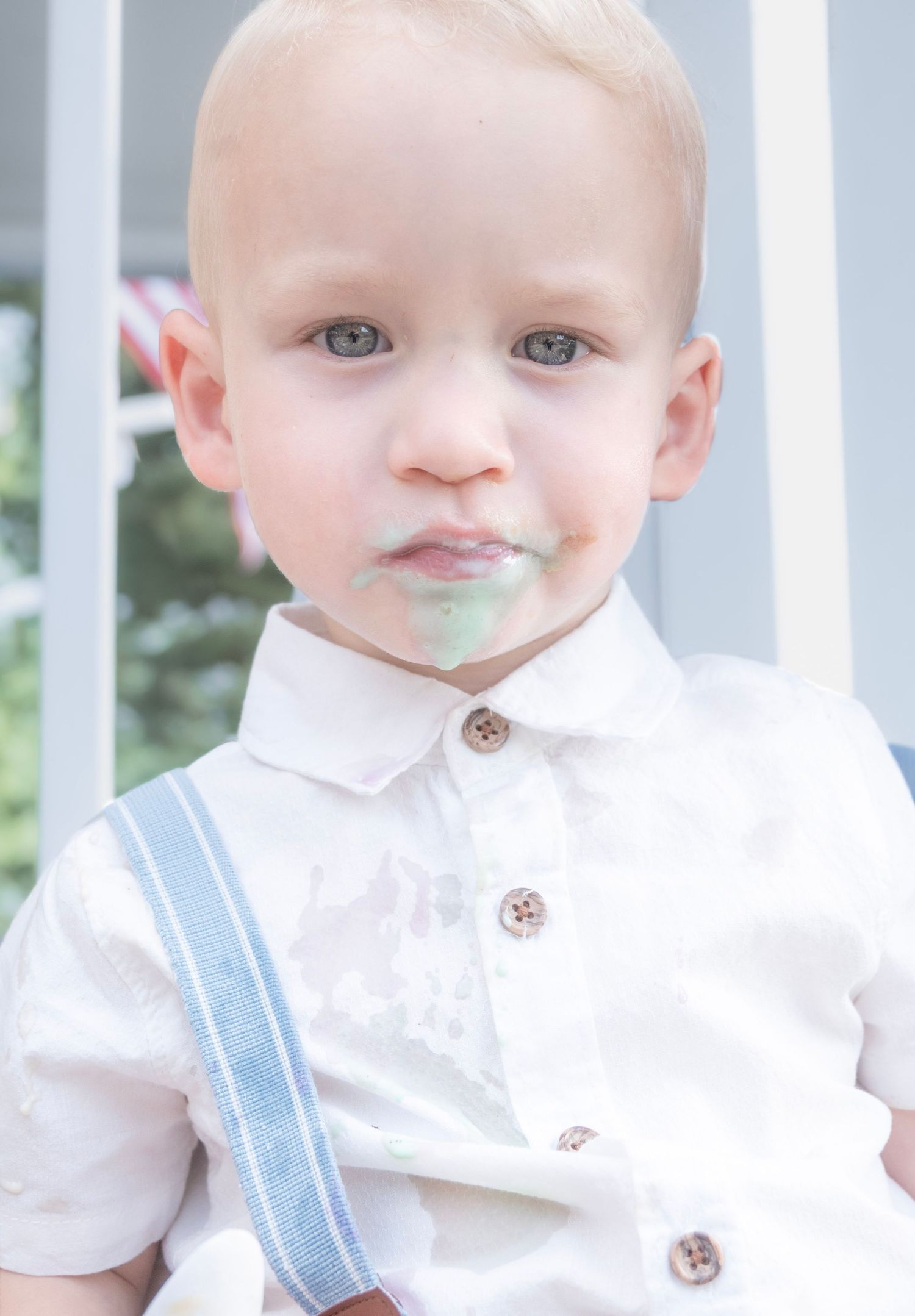 Adorable blonde toddler boy in white shirt and blue suspenders with frosting on his face at a birthday party.
