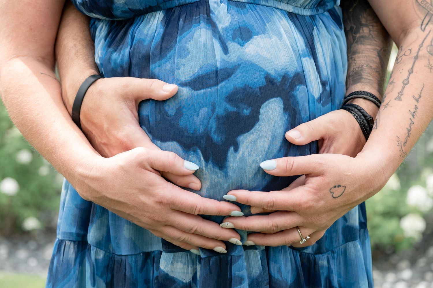 Couple forming heart shape with hands on pregnant woman's belly in blue floral dress outdoors.