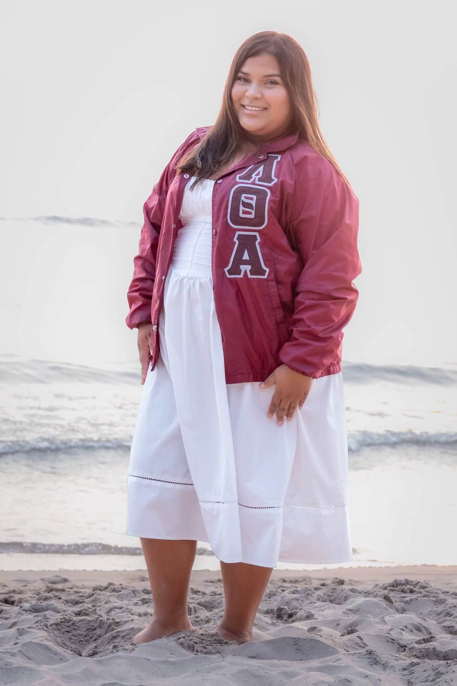 Young woman in white dress and maroon Greek letter jacket smiling on sandy beach near ocean.
