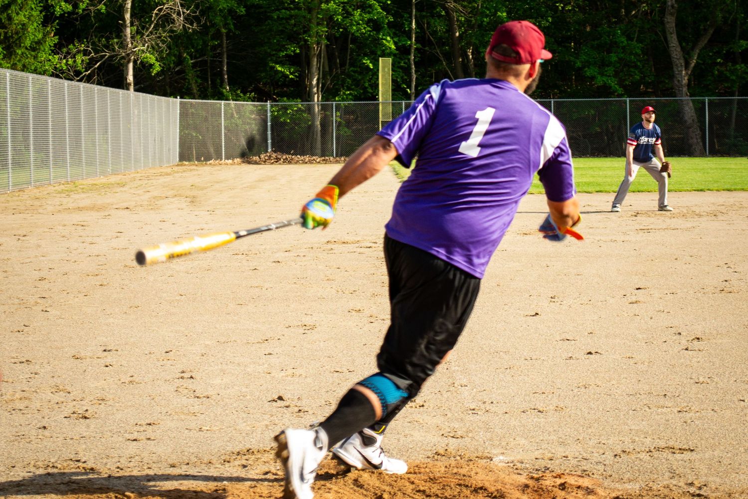 Softball batter in purple jersey number 1 swinging at a pitch during an outdoor recreational game.