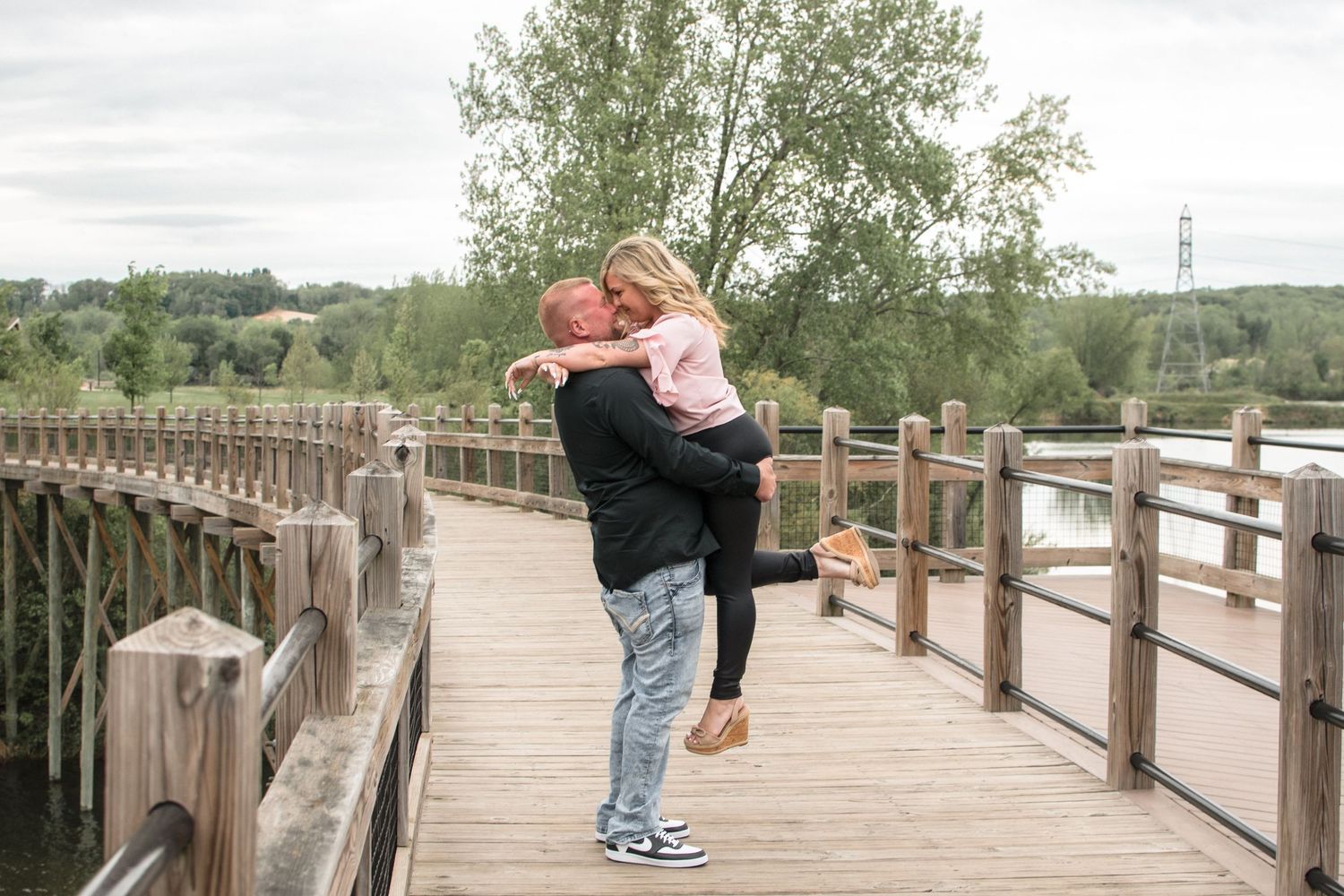 Couple sharing a romantic moment on a wooden bridge, man lifting woman as they kiss outdoors.