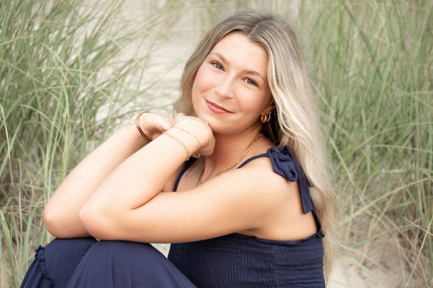 Smiling blonde woman in navy outfit sitting on sandy beach with tall grass, casual outdoor portrait photography.