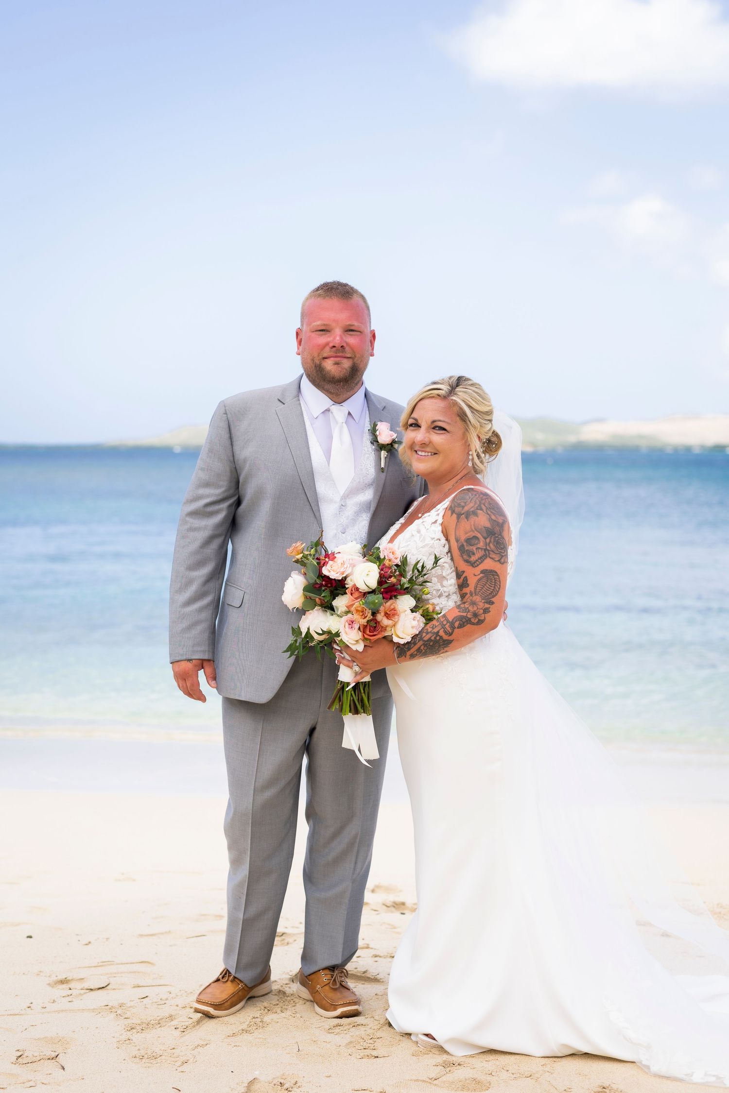 Bride and groom pose on a tropical beach, she holds a coral bouquet in a white gown, he wears a gray suit.