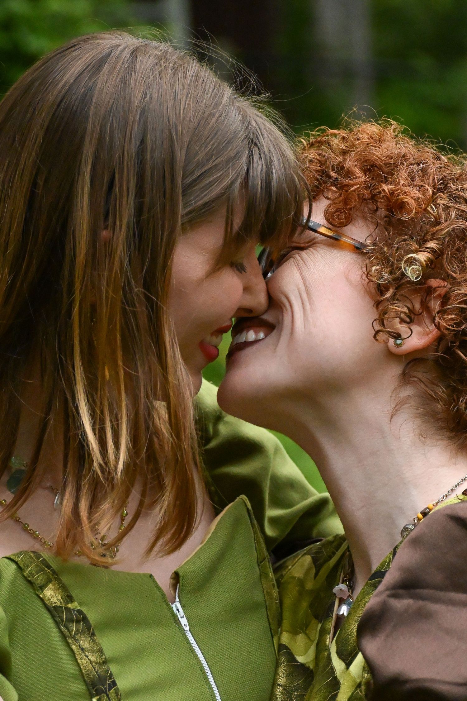 Two women smiling and leaning close together outdoors, one with straight brown hair and one with curly red hair.
