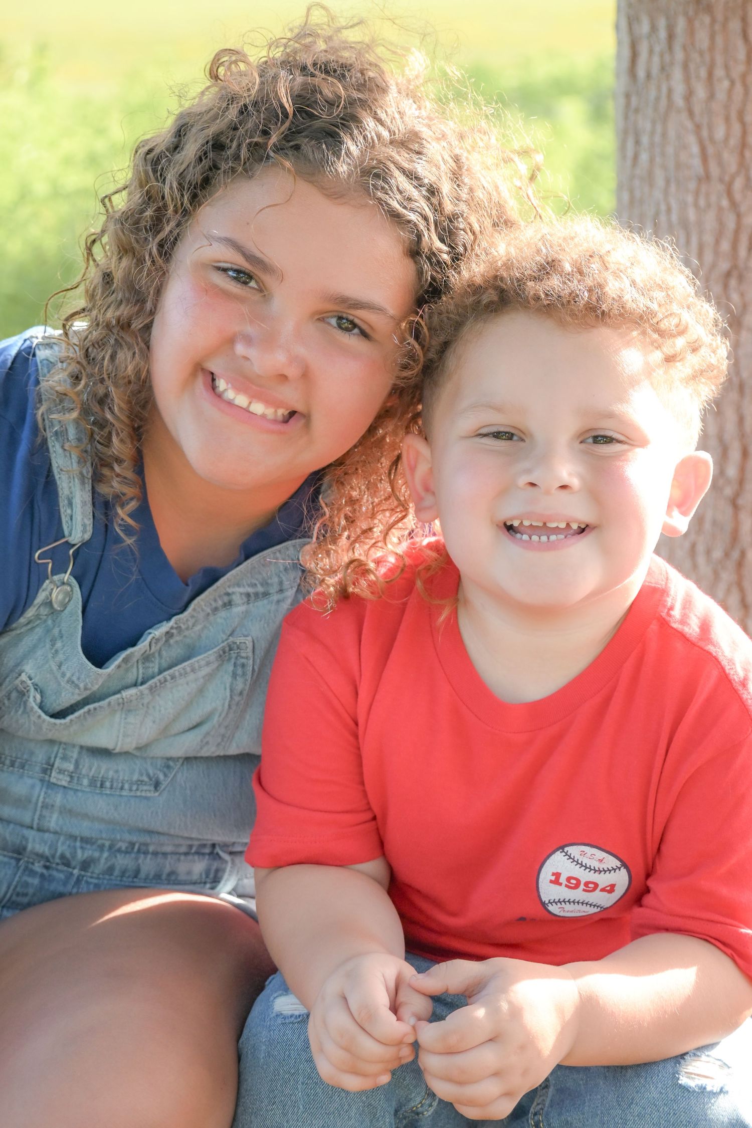 Smiling girl in denim overalls poses with young curly-haired boy in red baseball shirt outdoors.