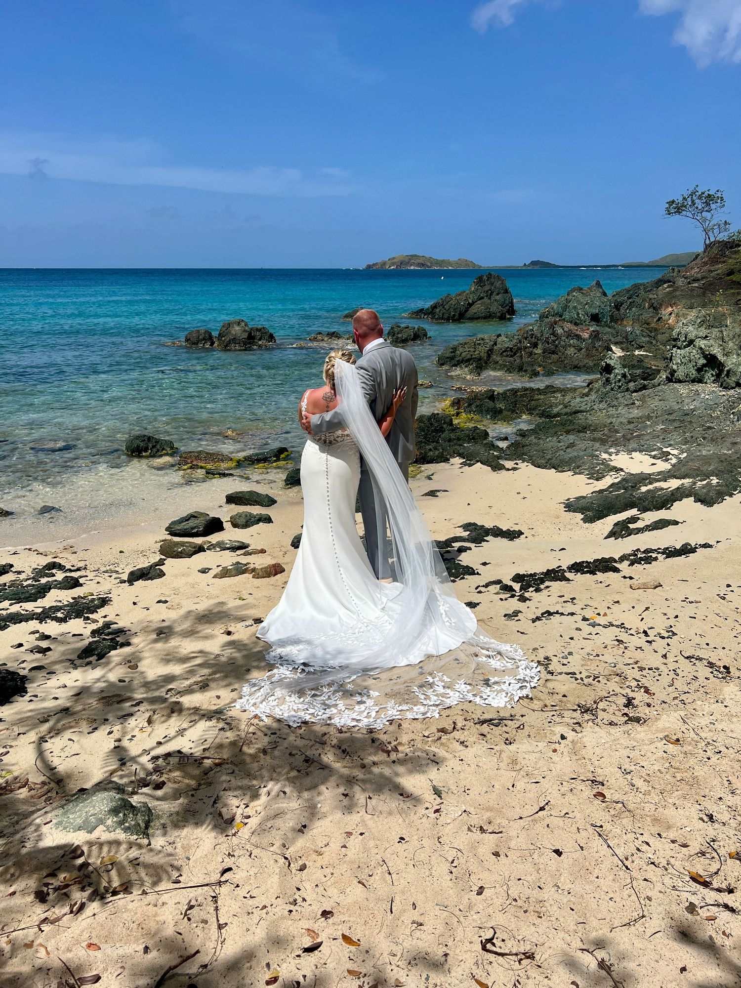 Newlywed couple kissing on a tropical beach with turquoise water and rocky shoreline on their wedding day.