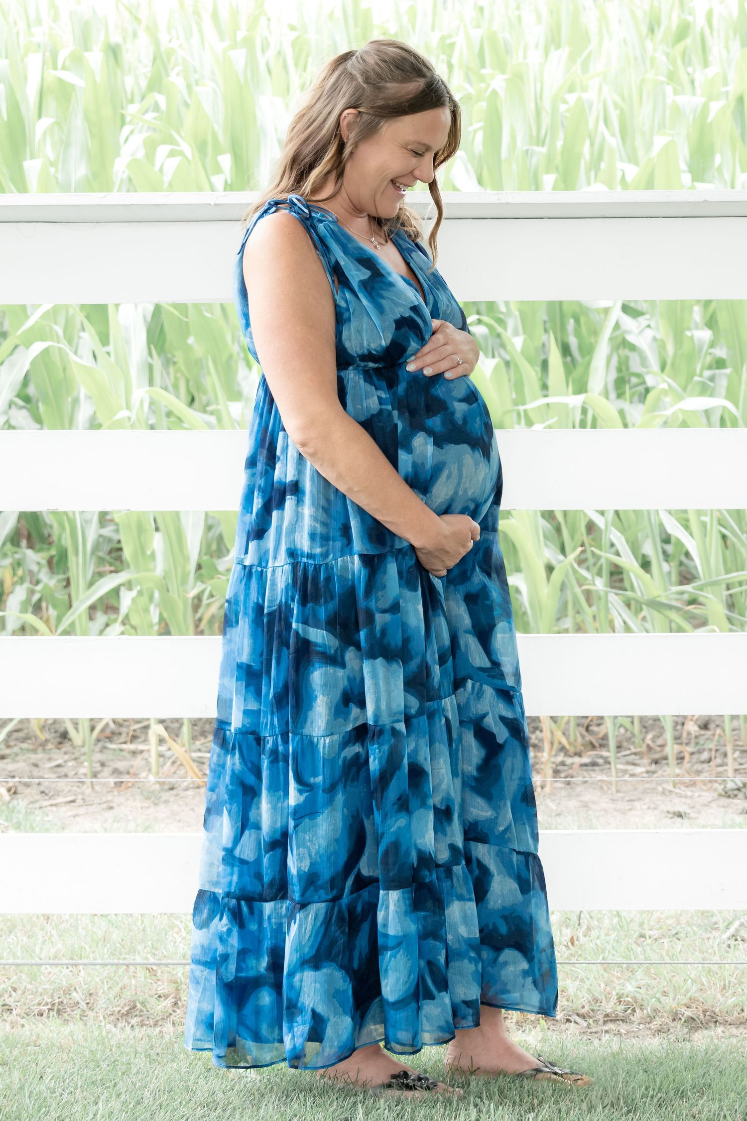 Pregnant woman in blue tie-dye maxi dress holding baby bump, smiling outdoors by white fence.