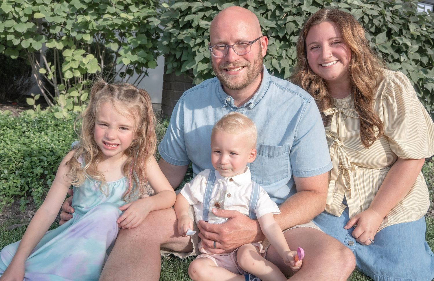 Family of four posing outdoors, parents with two young children smiling in front of lush green foliage.