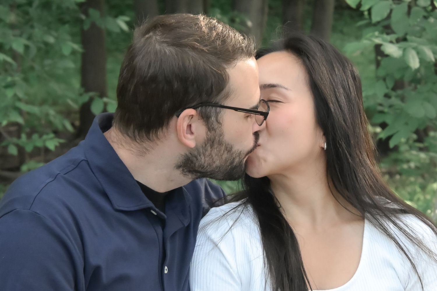 A couple sharing a romantic kiss outdoors surrounded by lush green trees in a natural setting.