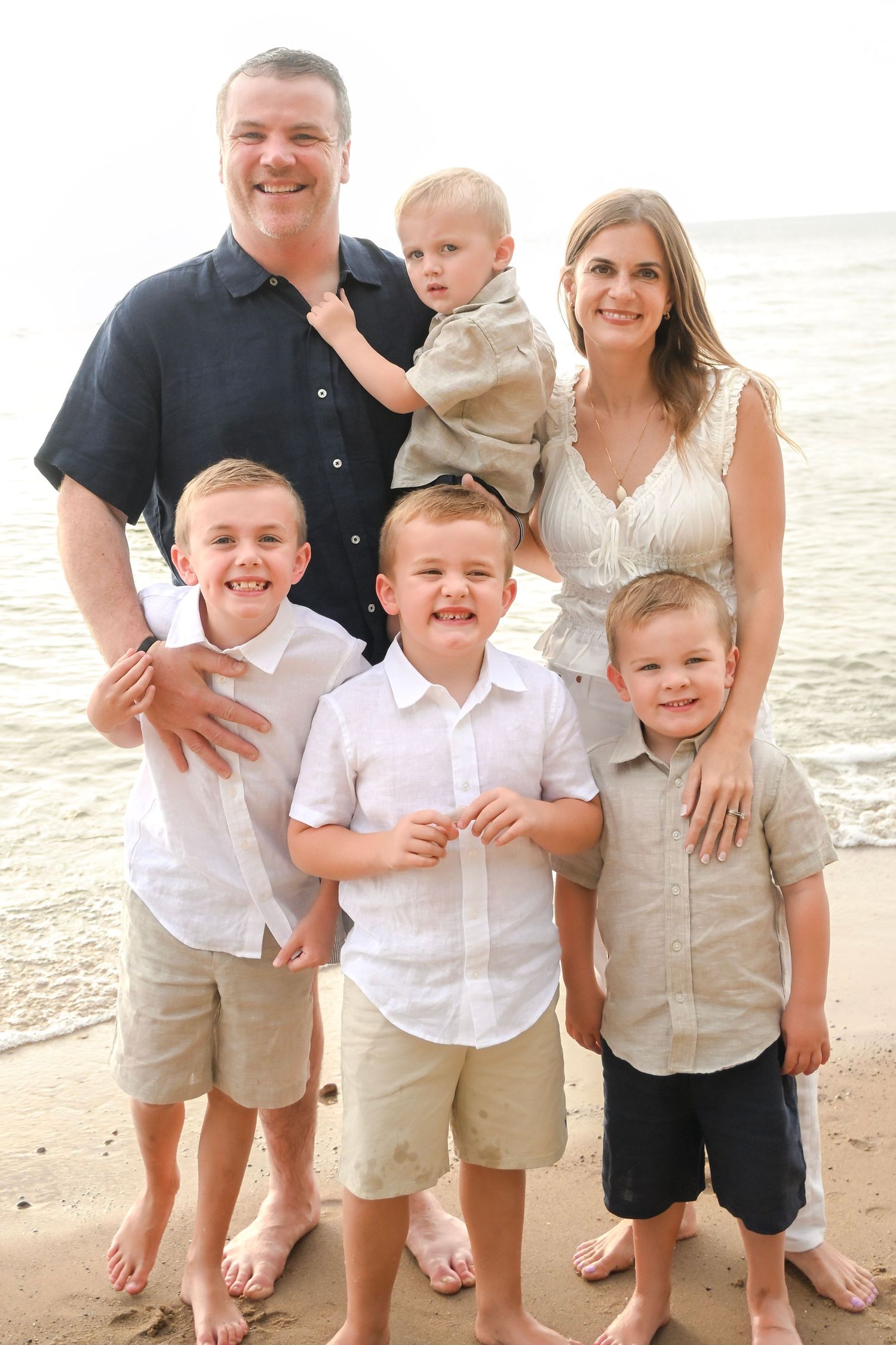 Family of six posing together on a sandy beach, parents with four young boys smiling in coordinated outfits.