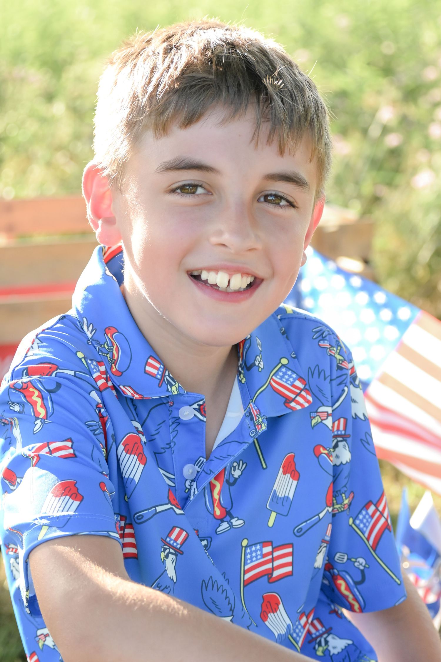 Smiling boy in patriotic blue shirt with American flag prints outdoors on a sunny day.