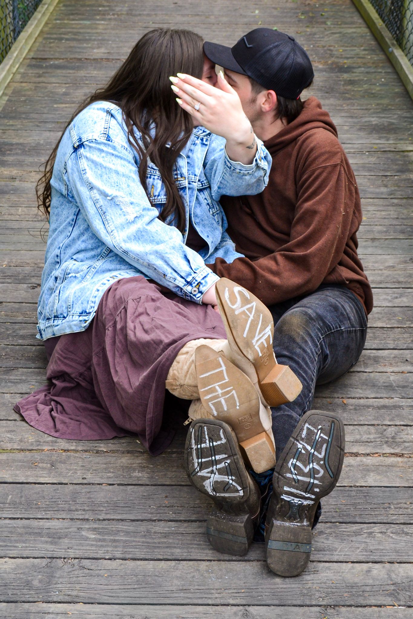 Newly engaged couple kissing on wooden bridge with save the date written on their shoes.