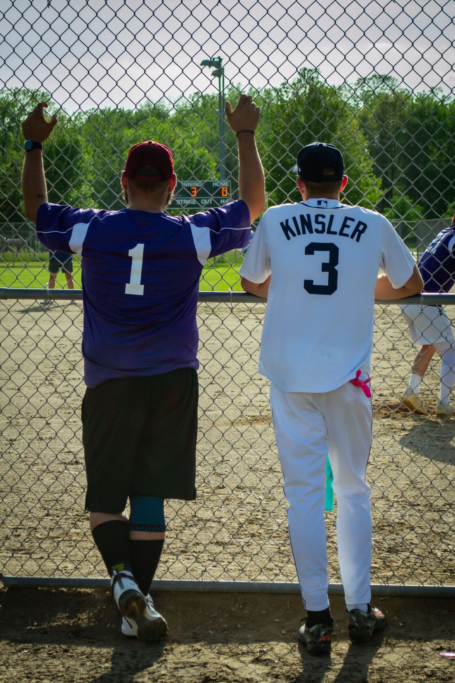 Two baseball fans wearing jerseys number 1 and Kinsler number 3 watching a game through a chain-link fence.