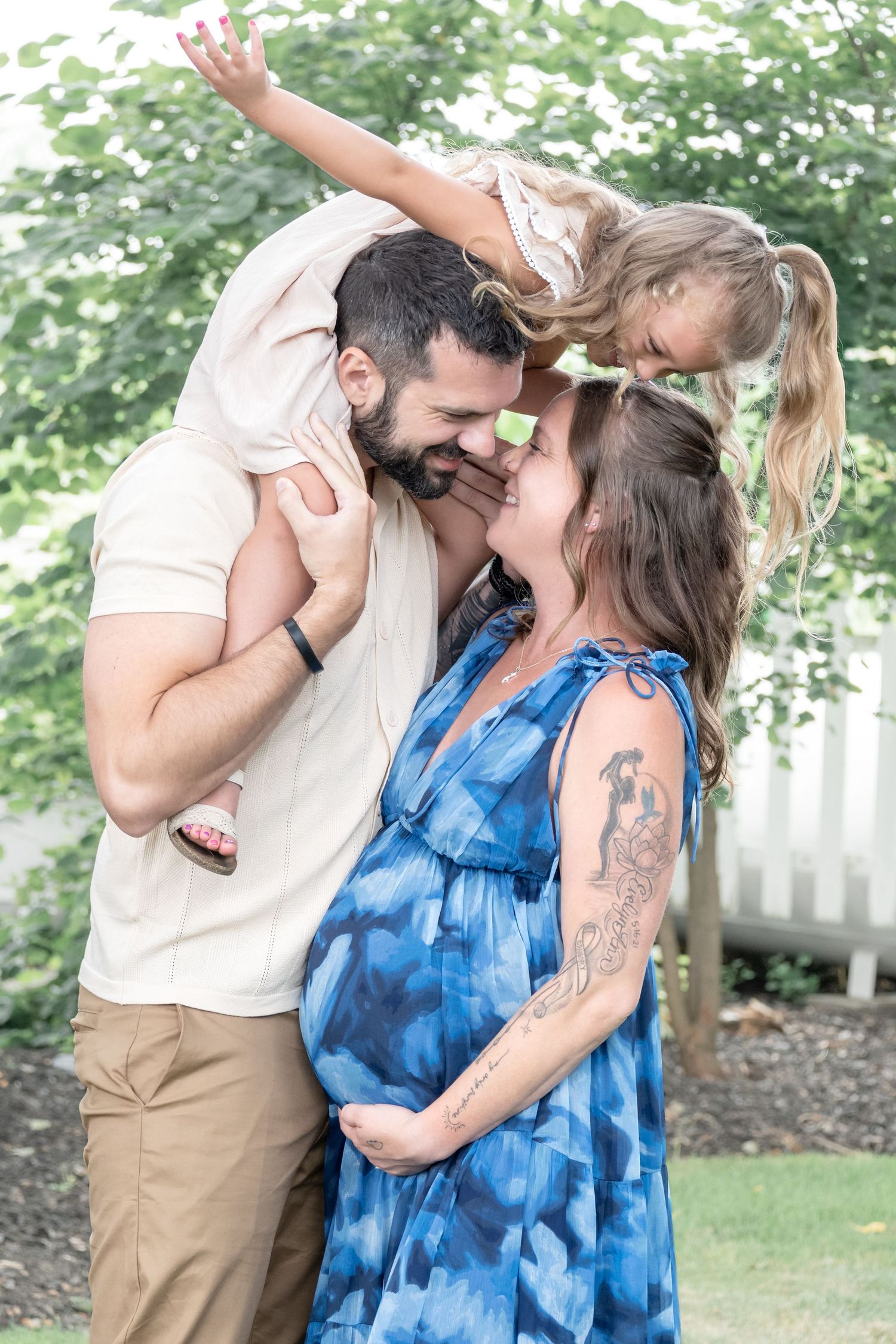 Pregnant woman in blue dress with bearded man and young girl on his shoulders sharing a joyful family moment outdoors.