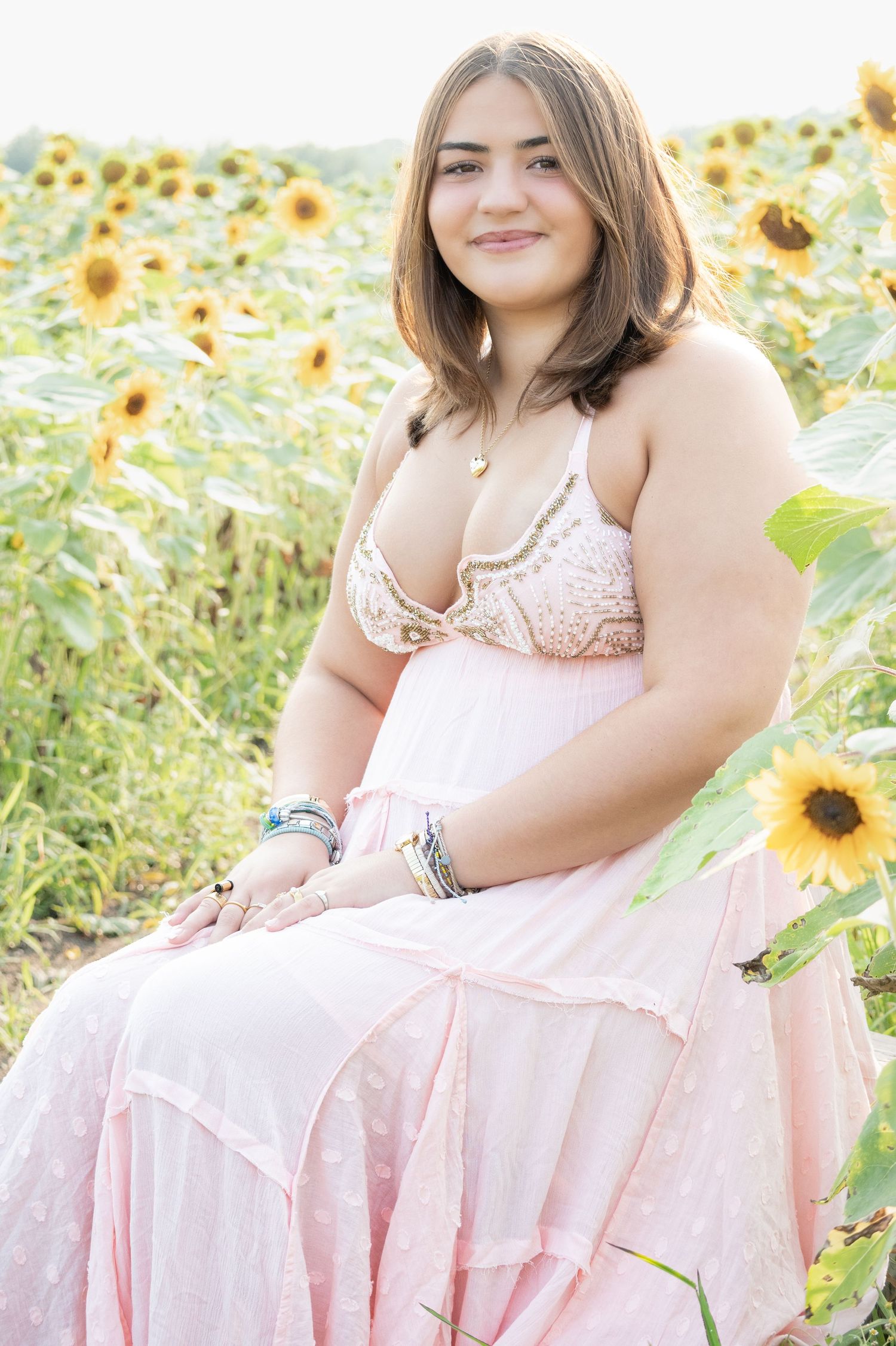 Young woman in pink dress sitting in sunflower field, smiling outdoors on a sunny day.
