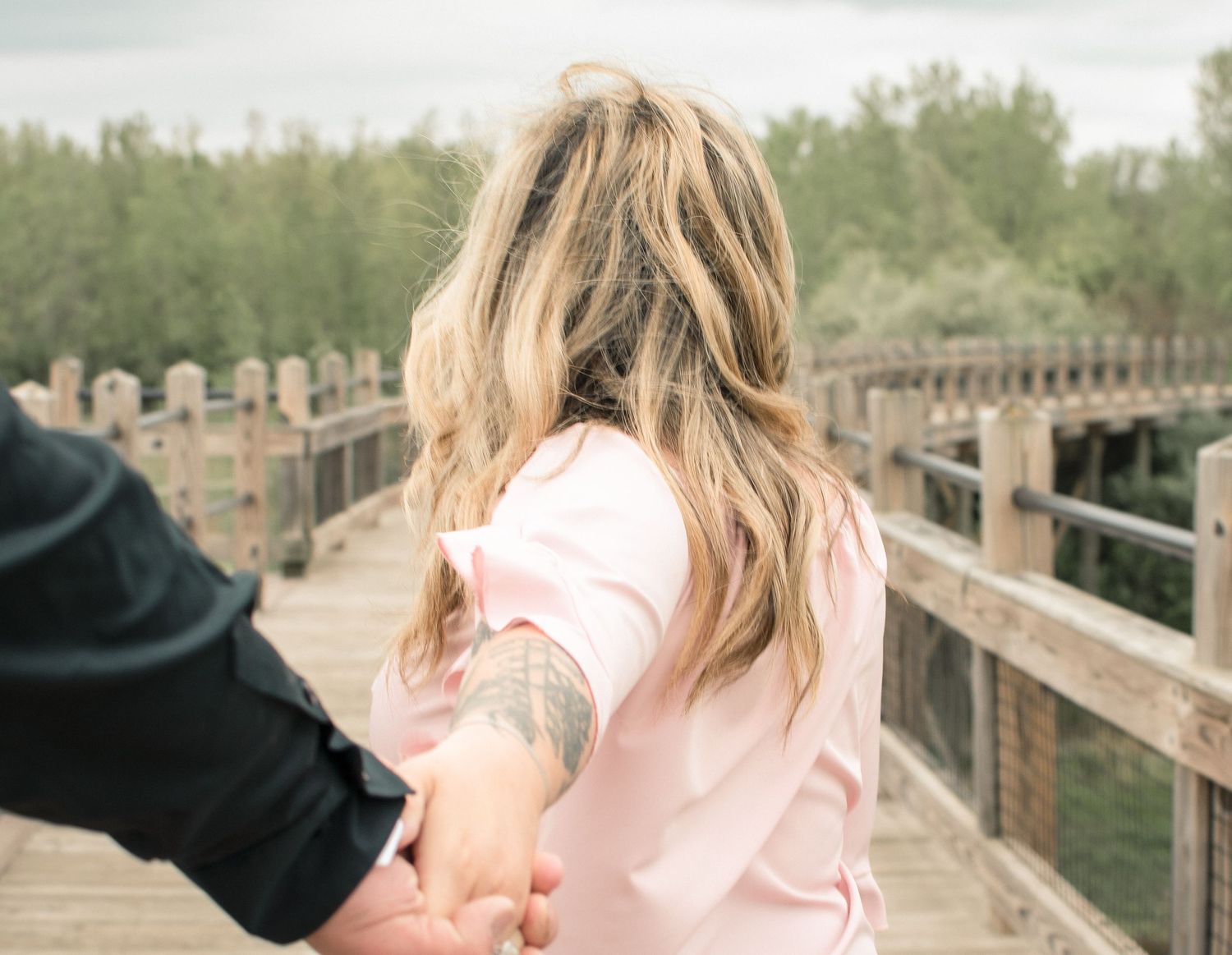 Woman with blonde hair holding a tattooed hand while walking on a wooden bridge outdoors.