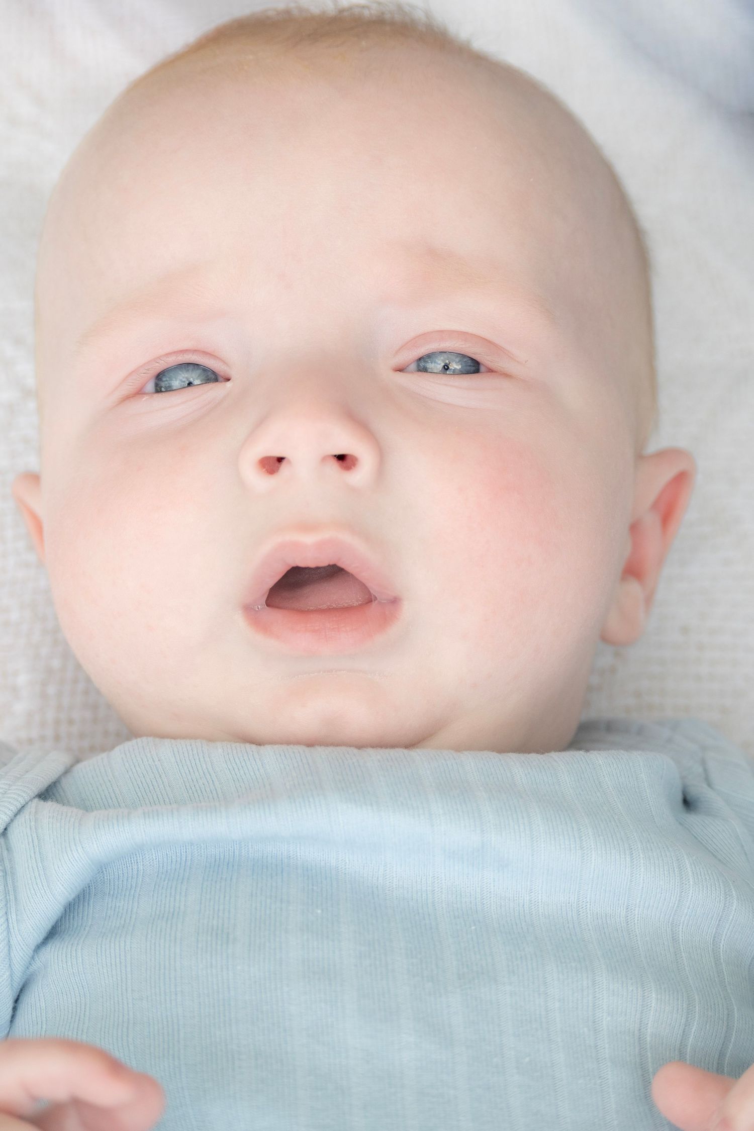 A cute baby with blue eyes and rosy cheeks lying on a white blanket wearing a light blue onesie.