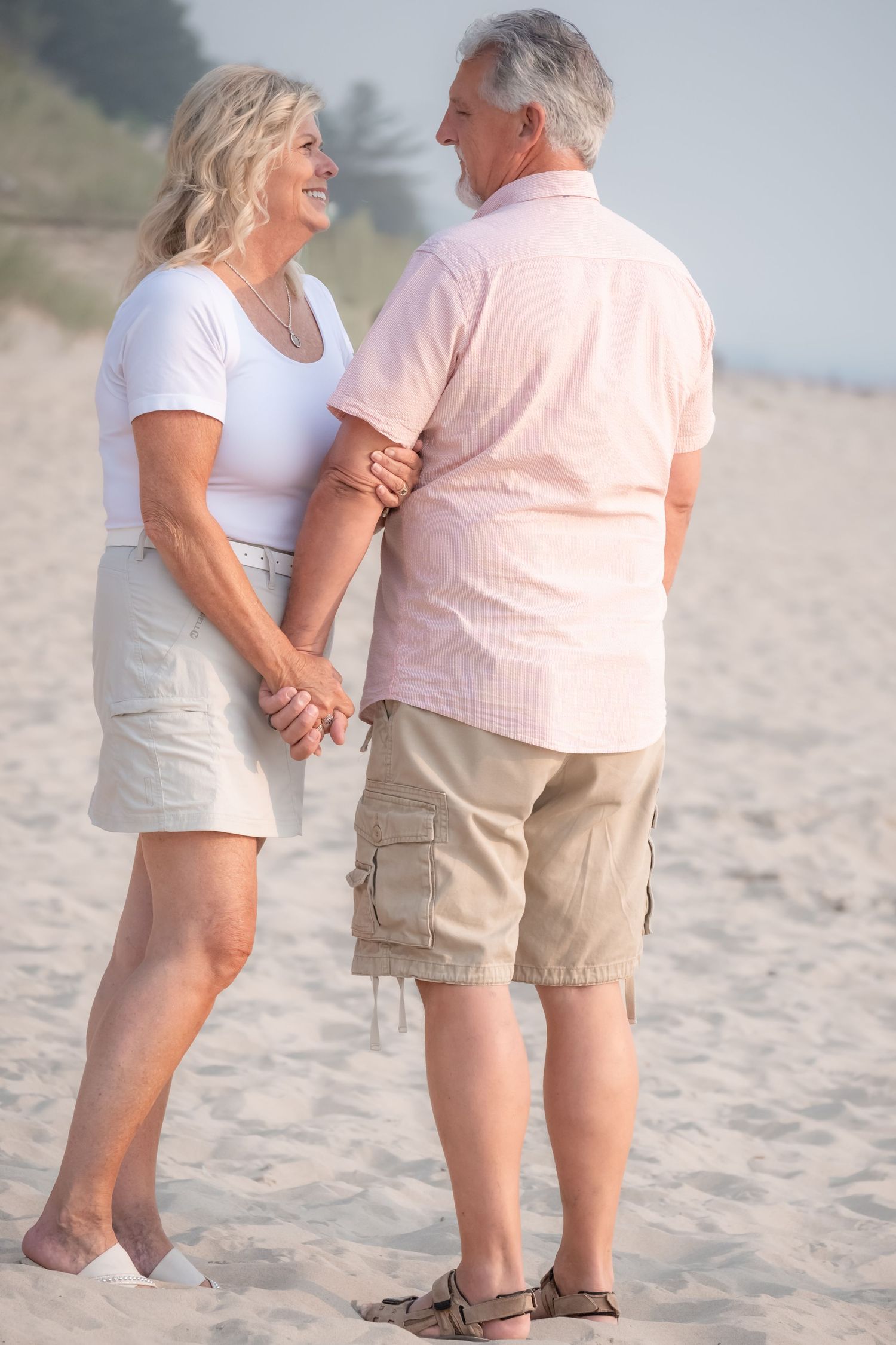 Smiling senior couple holding hands on a sandy beach, woman in white top and man in pink shirt enjoying a romantic walk.
