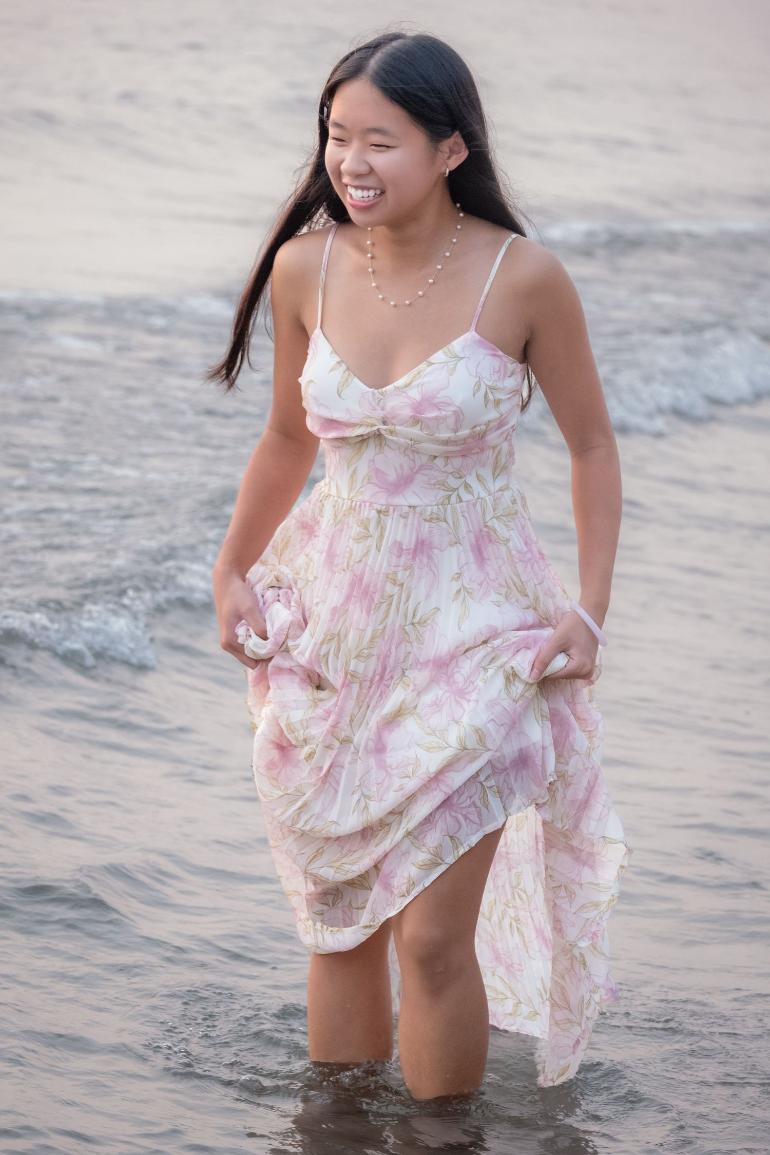 Smiling young Asian woman in floral sundress wading in ocean waves at beach, holding dress above water.