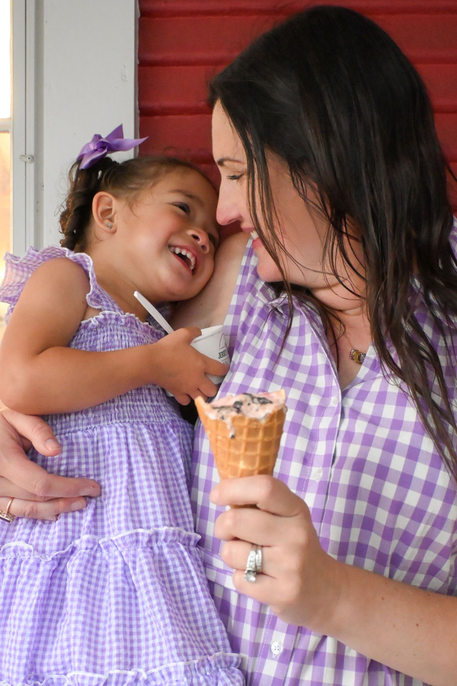 A laughing toddler girl in purple dress sharing a joyful moment with her mother holding an ice cream cone.