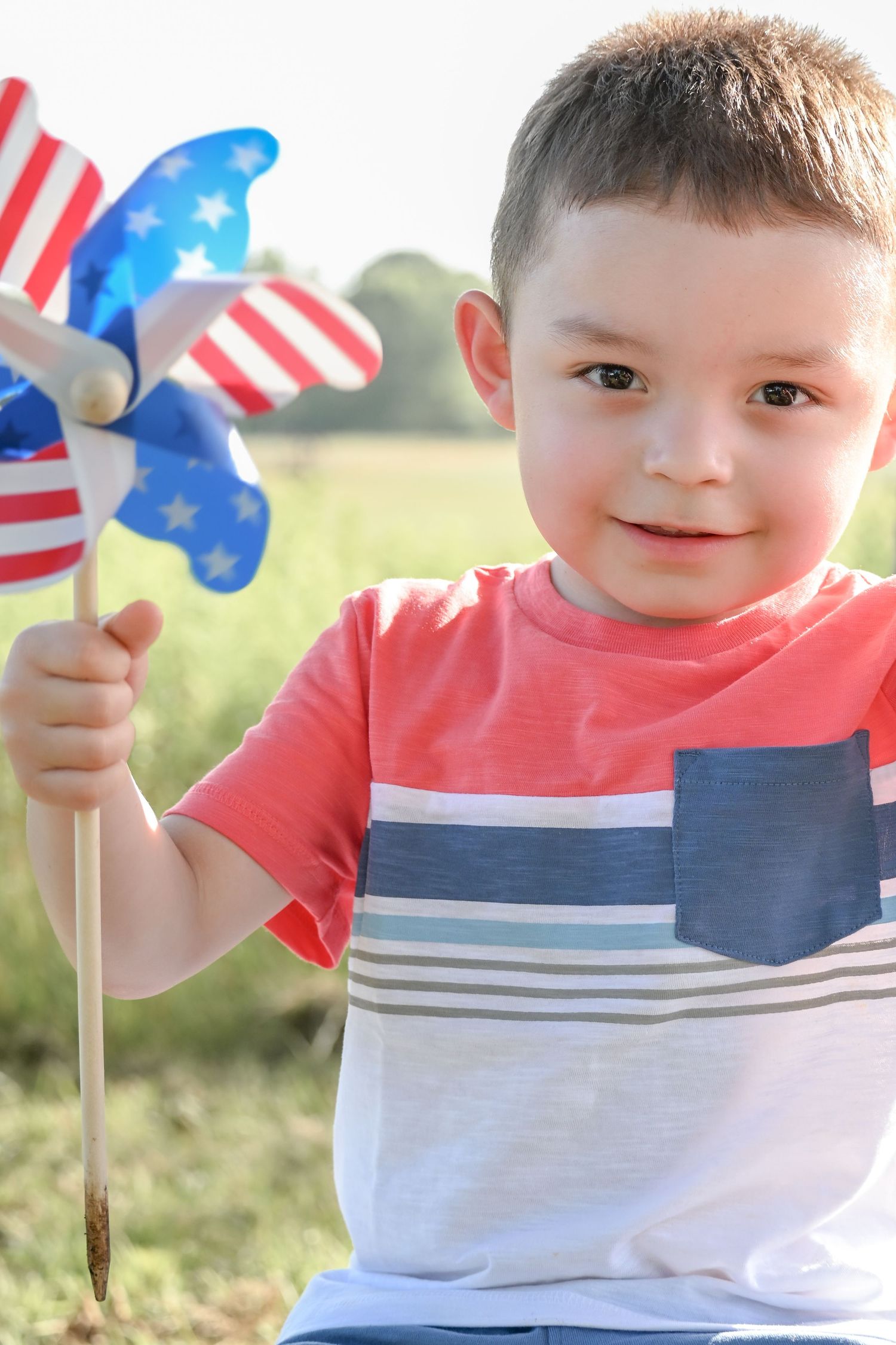 Young boy smiling outdoors holding an American flag patriotic pinwheel on a sunny day.