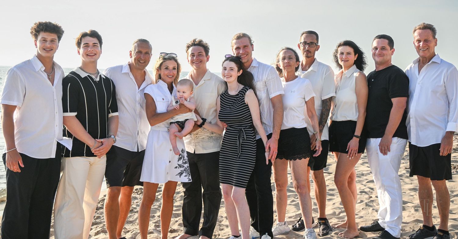 Large family group portrait smiling together on a sunny beach, dressed in white and black outfits.