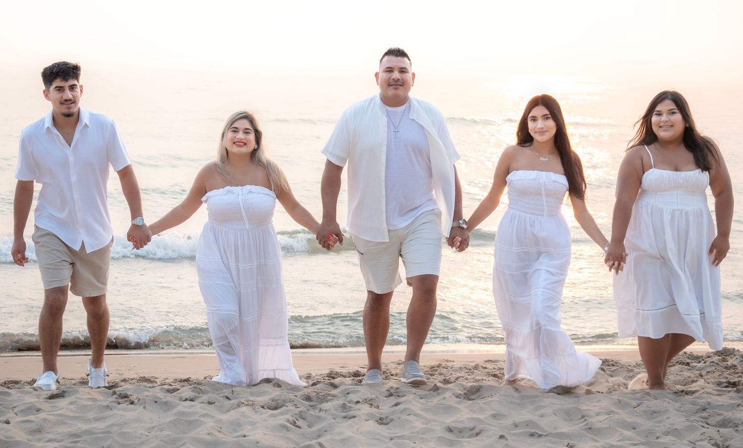Family of five in white outfits holding hands on a sandy beach at sunset.