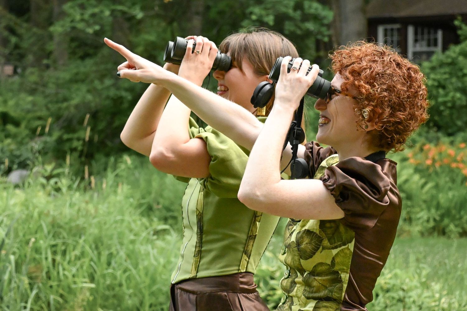 Two women using binoculars for birdwatching outdoors in a lush green forest setting.