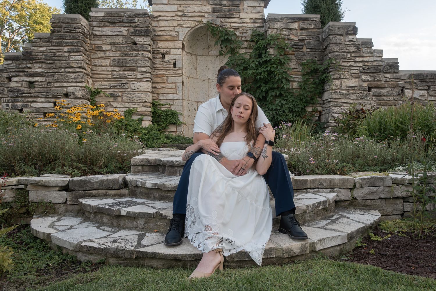 Couple in wedding attire sitting on stone steps surrounded by rustic stone ruins and lush greenery.
