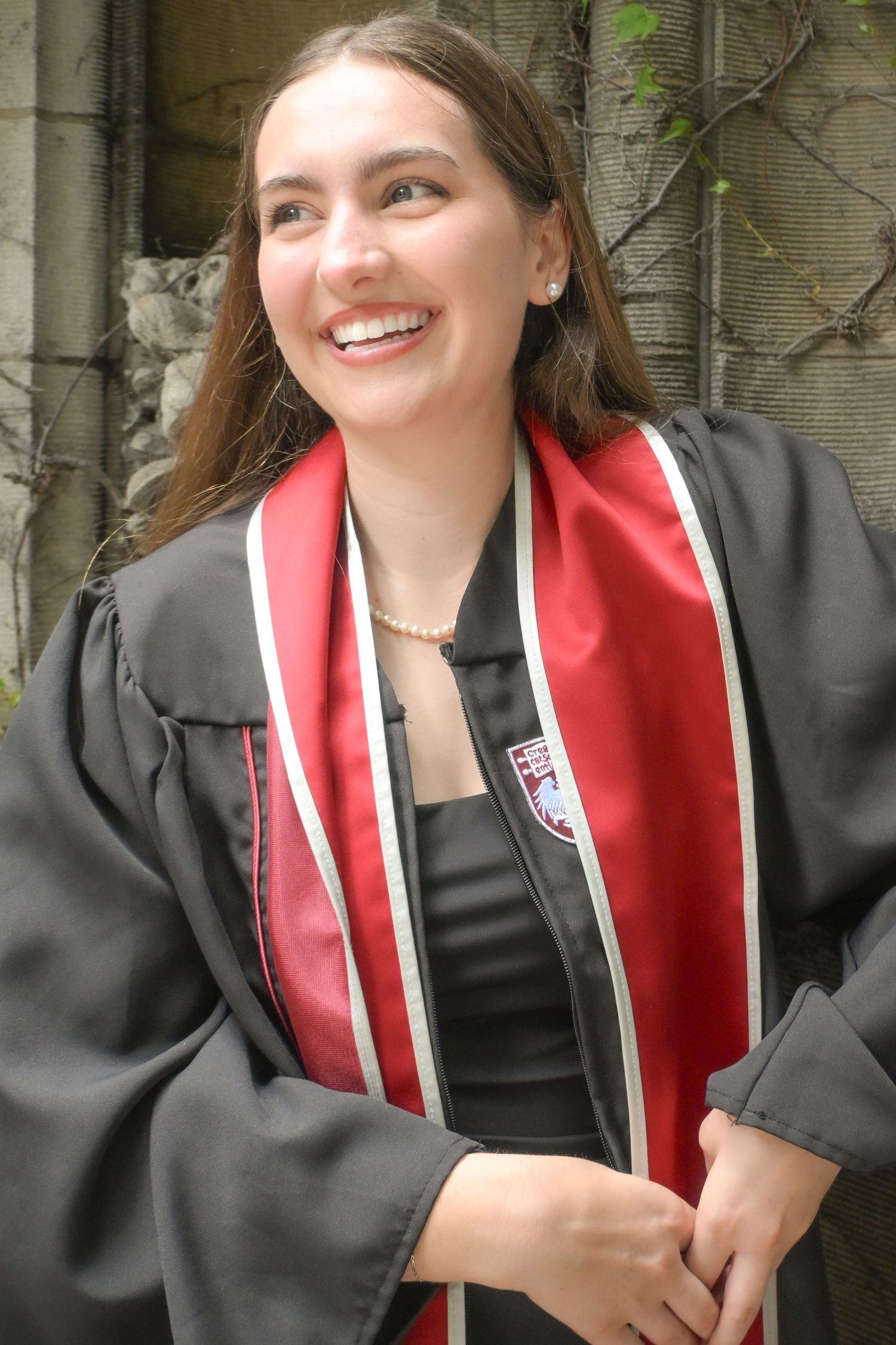 Smiling female college graduate wearing black graduation gown and red stole with school crest outdoors.